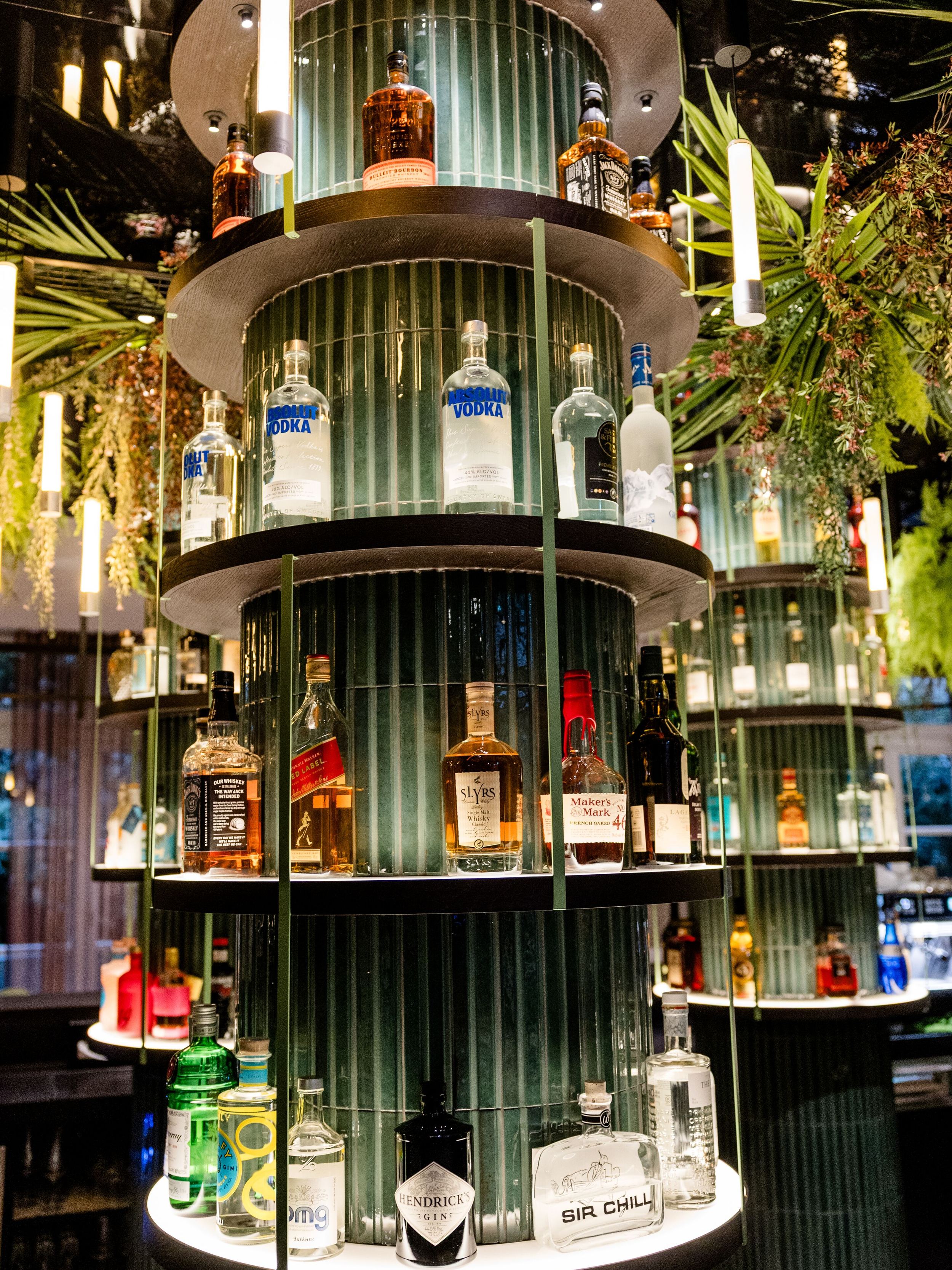 Hotel Frankfurt-Hauptbahnhof Motel One mehrstöckiges beleuchtetes Regal mit verschiedenen Spirituosenflaschen, umgeben von grünen Pflanzen, in stilvoller Bar-Atmosphäre.