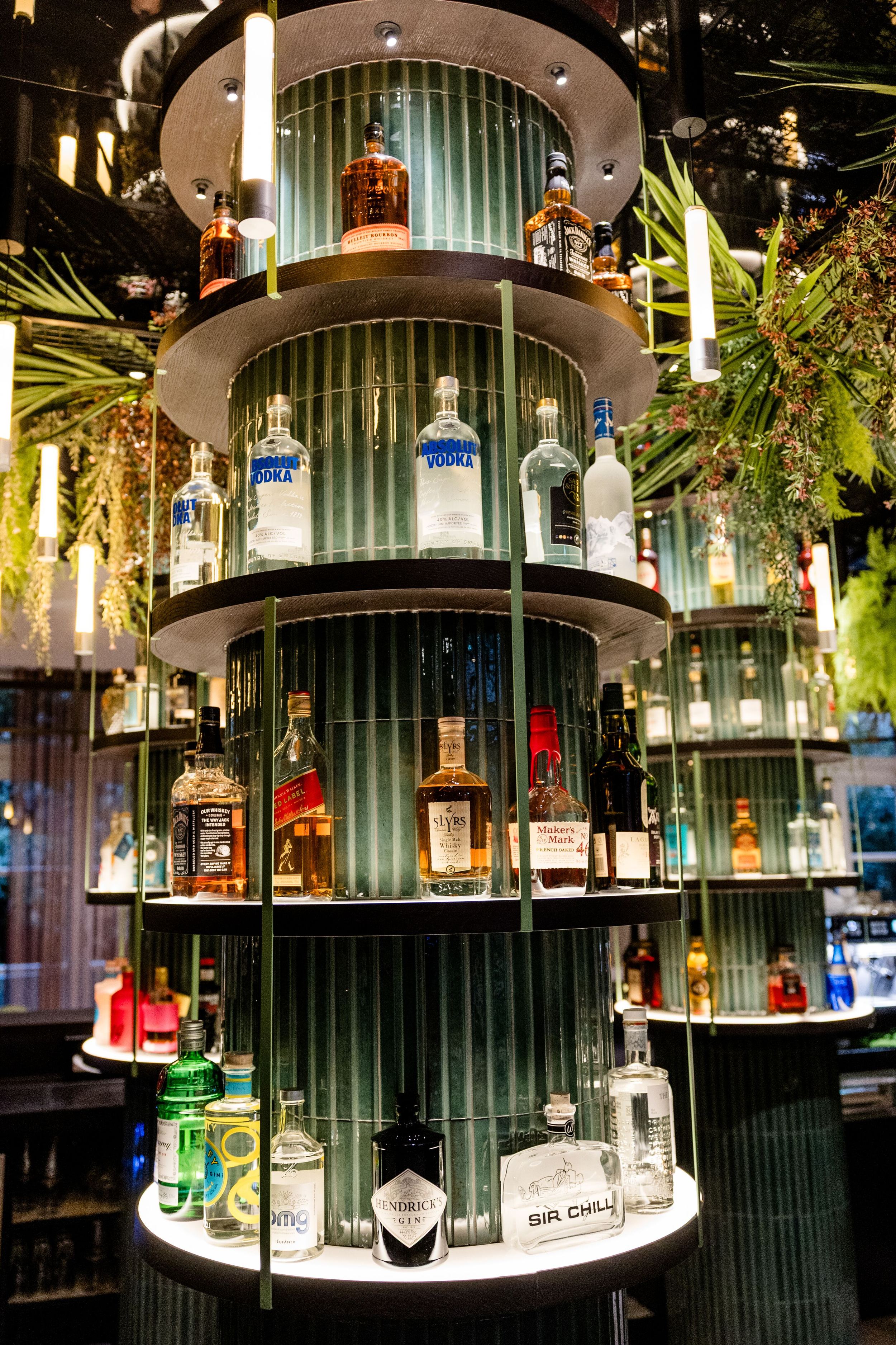 Hotel Frankfurt-Hauptbahnhof Motel One mehrstöckiges beleuchtetes Regal mit verschiedenen Spirituosenflaschen, umgeben von grünen Pflanzen, in stilvoller Bar-Atmosphäre.