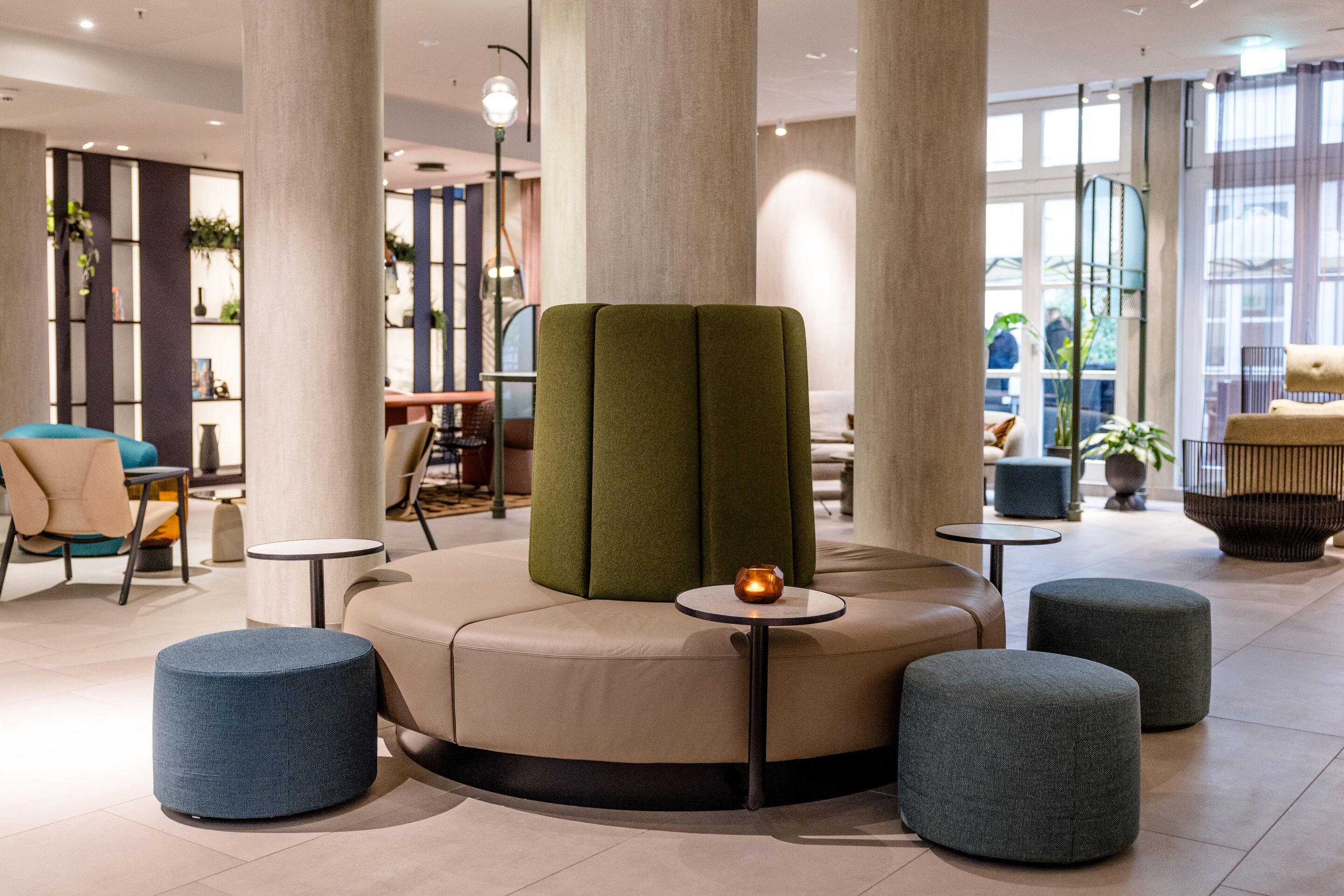 Hotel Frankfurt-Hauptbahnhof Motel One Lounge mit runder Sitzbank um Säule, bequemen Hockern und kleinem Tisch, warmes Ambiente mit natürlichem Licht und stilvoller Einrichtung.
