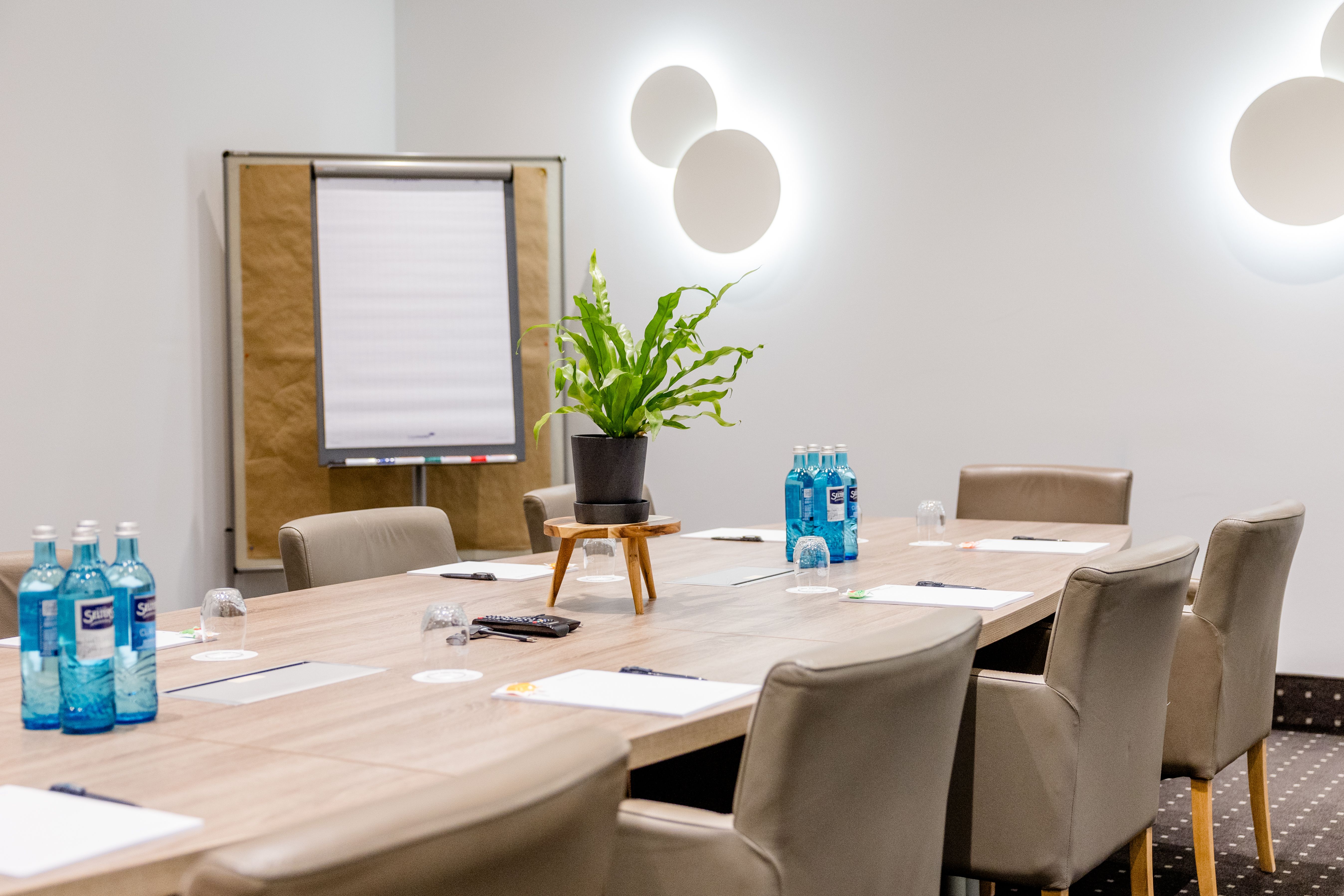 Moderner Konferenzraum mit großem Holztisch, bequemen Stühlen, Flipchart, Wasserflaschen und Notizblöcken, beleuchtet durch runde Wandlampen, Pflanzen als Dekoration. Ideal für Meetings oder Geschäftstreffen.