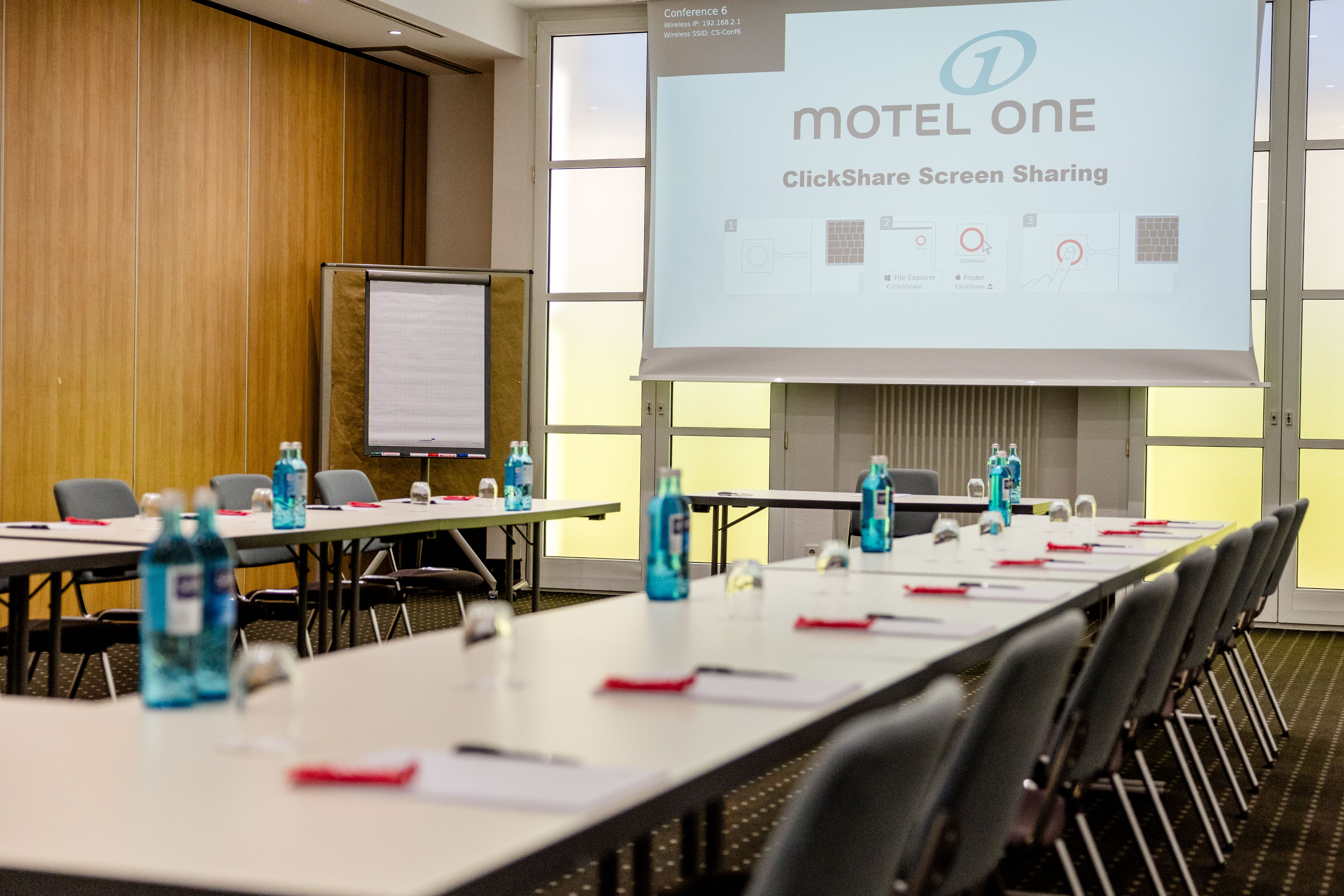 Konferenzraum mit schlanken Tischen, Stühlen und vorbereitetem Sitzplatz mit Wasserflaschen, Gläsern und Schreibmaterialien, Präsentation auf Leinwand mit Motel One-Logo und Screen-Sharing-Funktion. Helle Fenster sorgen für Tageslicht.