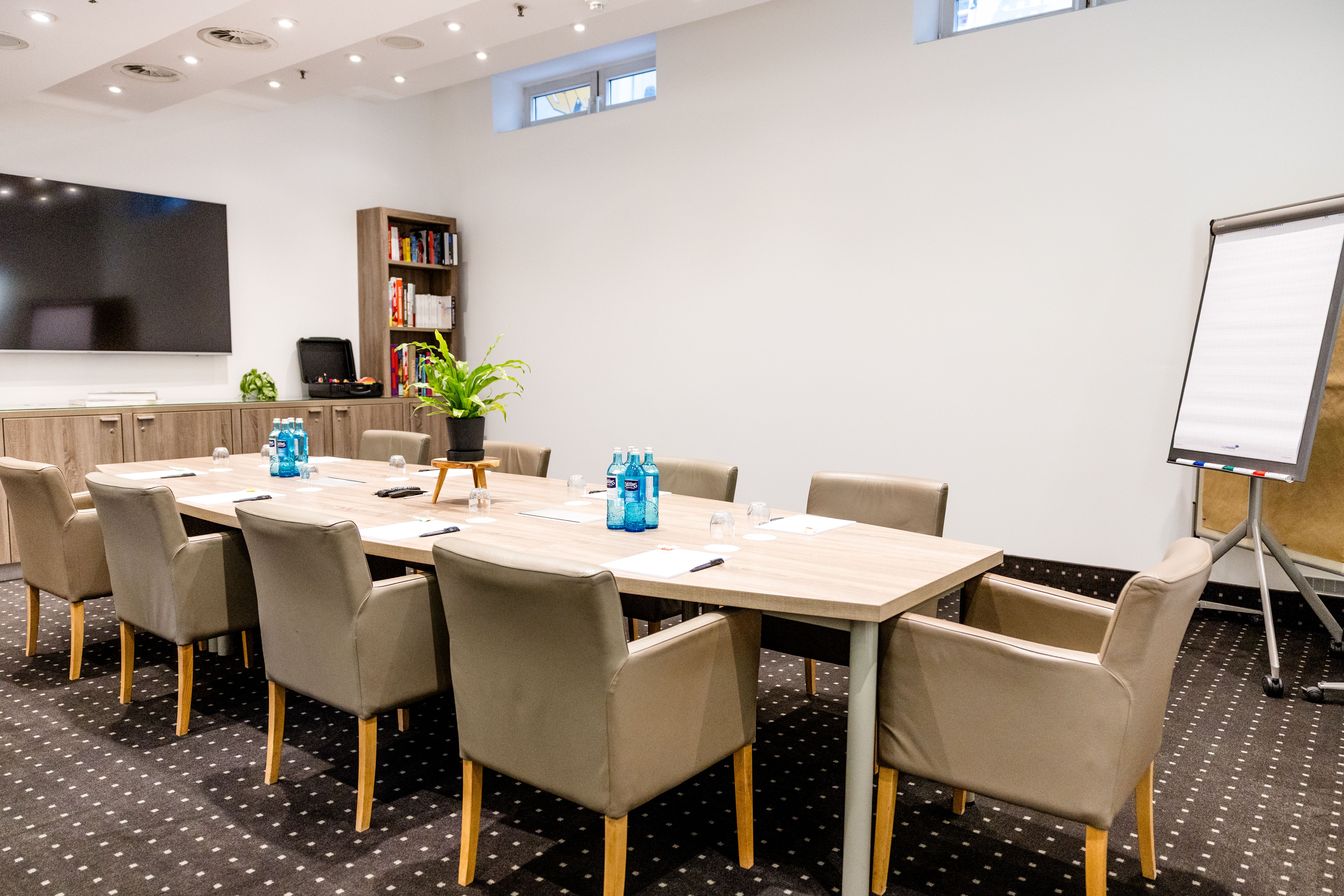 Moderner, heller Konferenzraum mit großem Tisch, komfortablen Stühlen, einem Flipchart und einem großen Fernseher, ideal für Meetings und Geschäftstreffen.