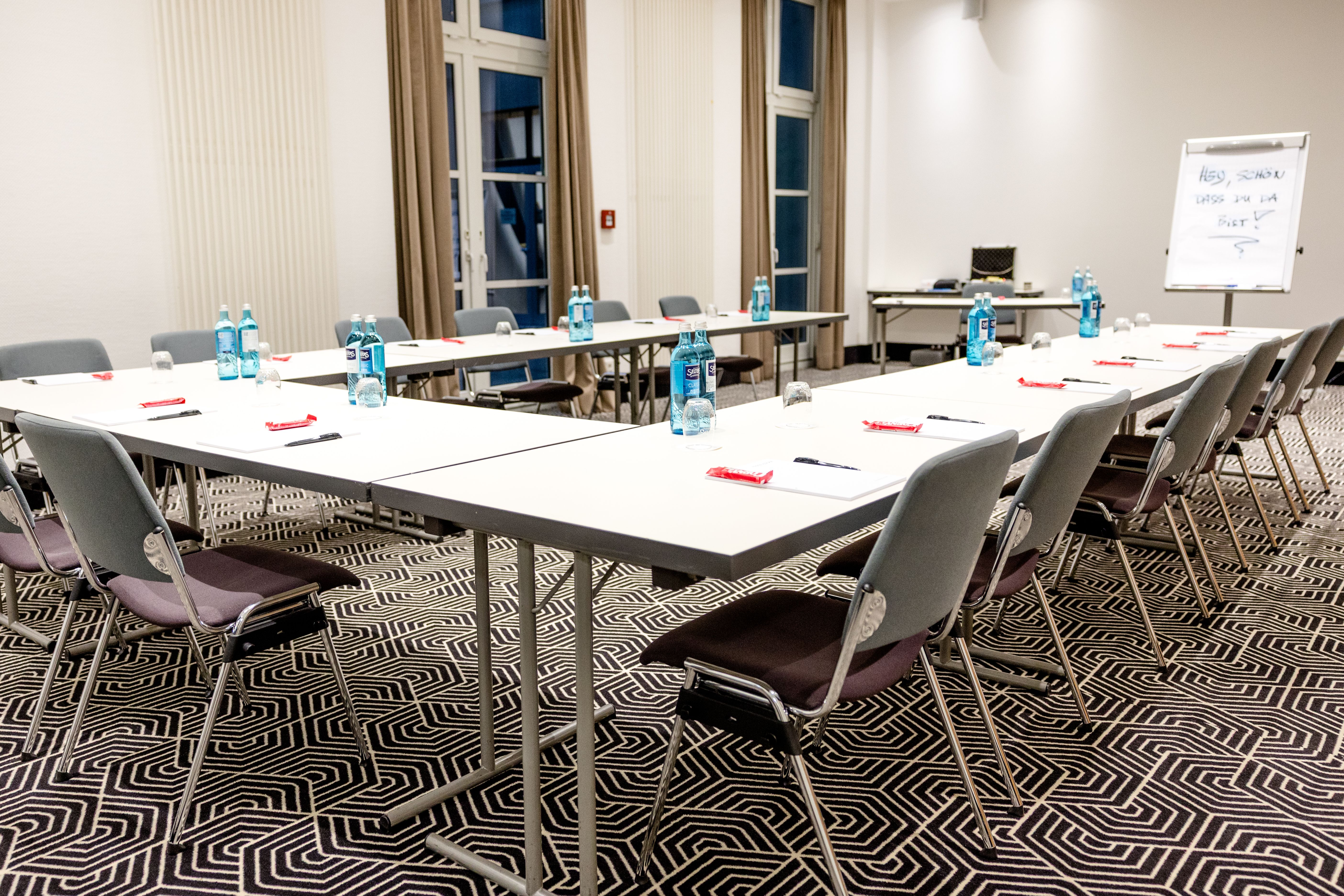 Konferenzraum mit U-förmig angeordneten Tischen, bestückt mit Stühlen, Wasserflaschen, Notizblöcken und Stiften, für Geschäftstreffen oder Seminare vorbereitet. Große Fenster sorgen für Tageslicht.