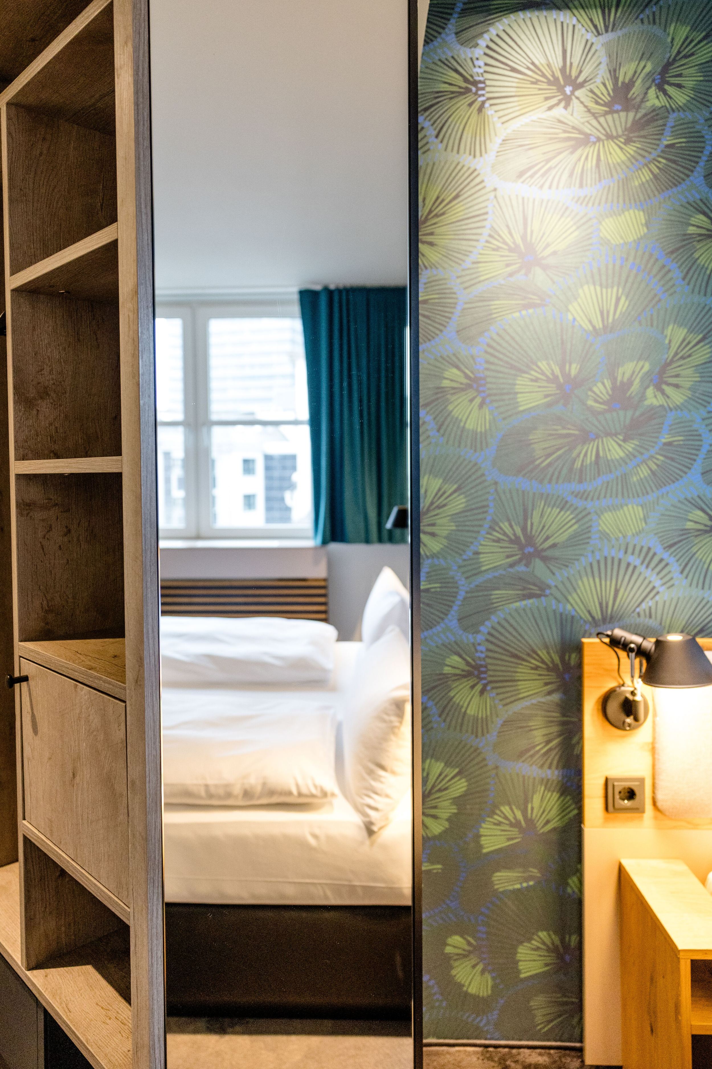 Designhotel Frankfurt Hauptbahnhof Motel One Blick in Hotelzimmer mit hellem Bett, grün-blau gemusterter Wand und indirekter Beleuchtung am Bettkopfbrett.