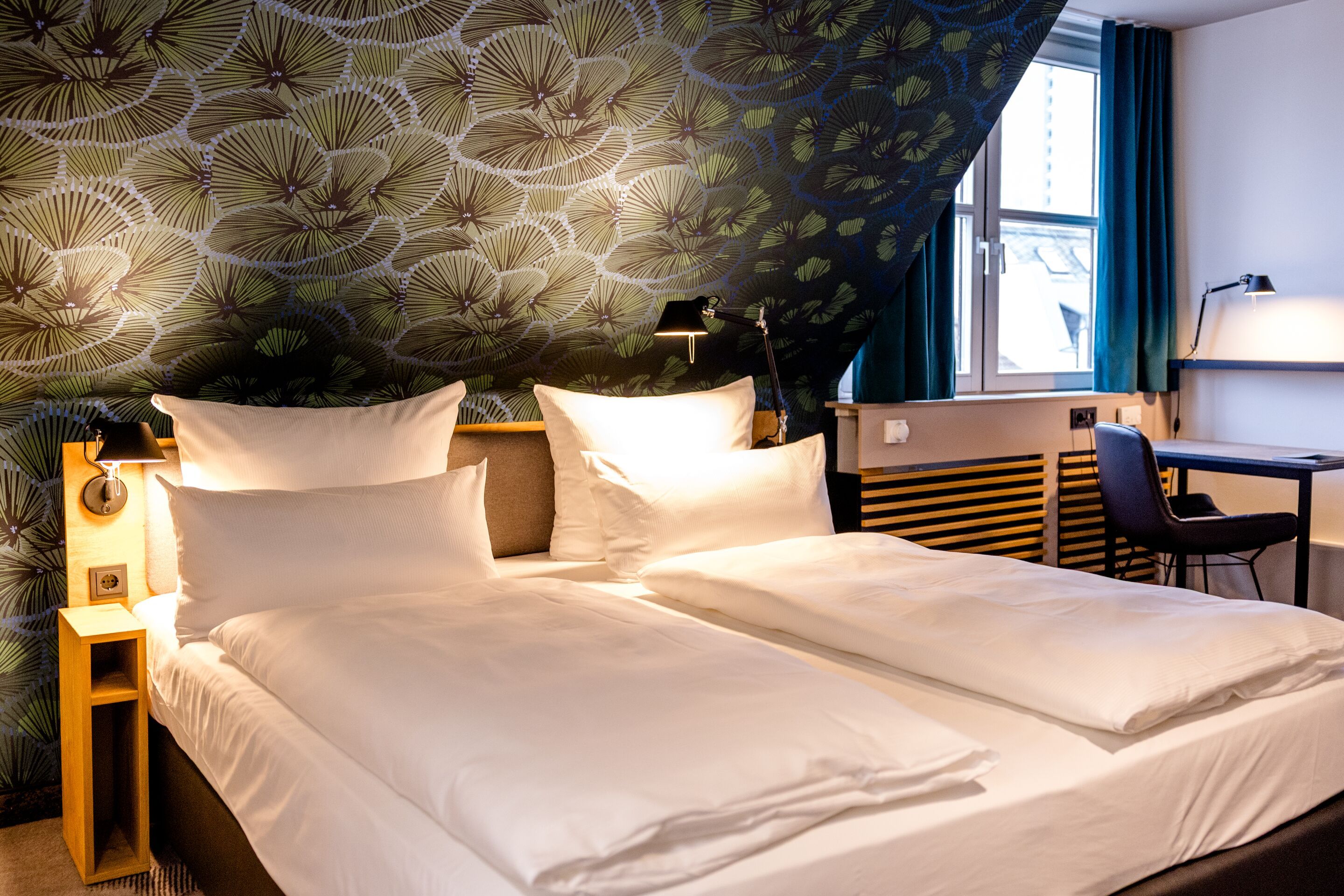 Hotel Frankfurt Hauptbahnhof Motel One Doppelzimmer mit zwei Einzelbetten, floral gemusterter Wand, weicher Beleuchtung und Arbeitsplatz am Fenster für erholsamen Komfort und produktives Arbeiten.