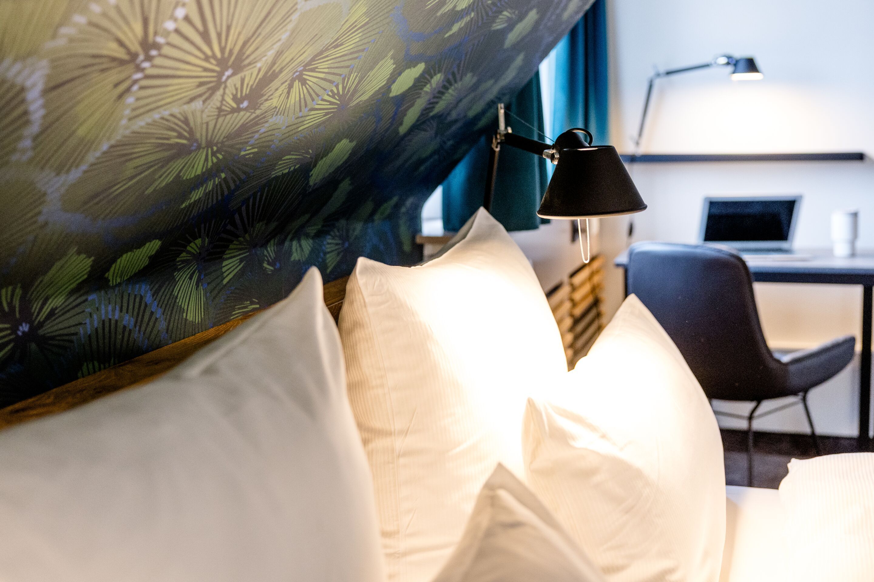 Hotel Frankfurt Hauptbahnhof Motel One Zimmer mit gemütlichem Bett, mehreren weißen Kissen und einer Tapete mit Blättermuster. Im Hintergrund ein Schreibtisch mit Stuhl und Arbeitsplatzbeleuchtung.