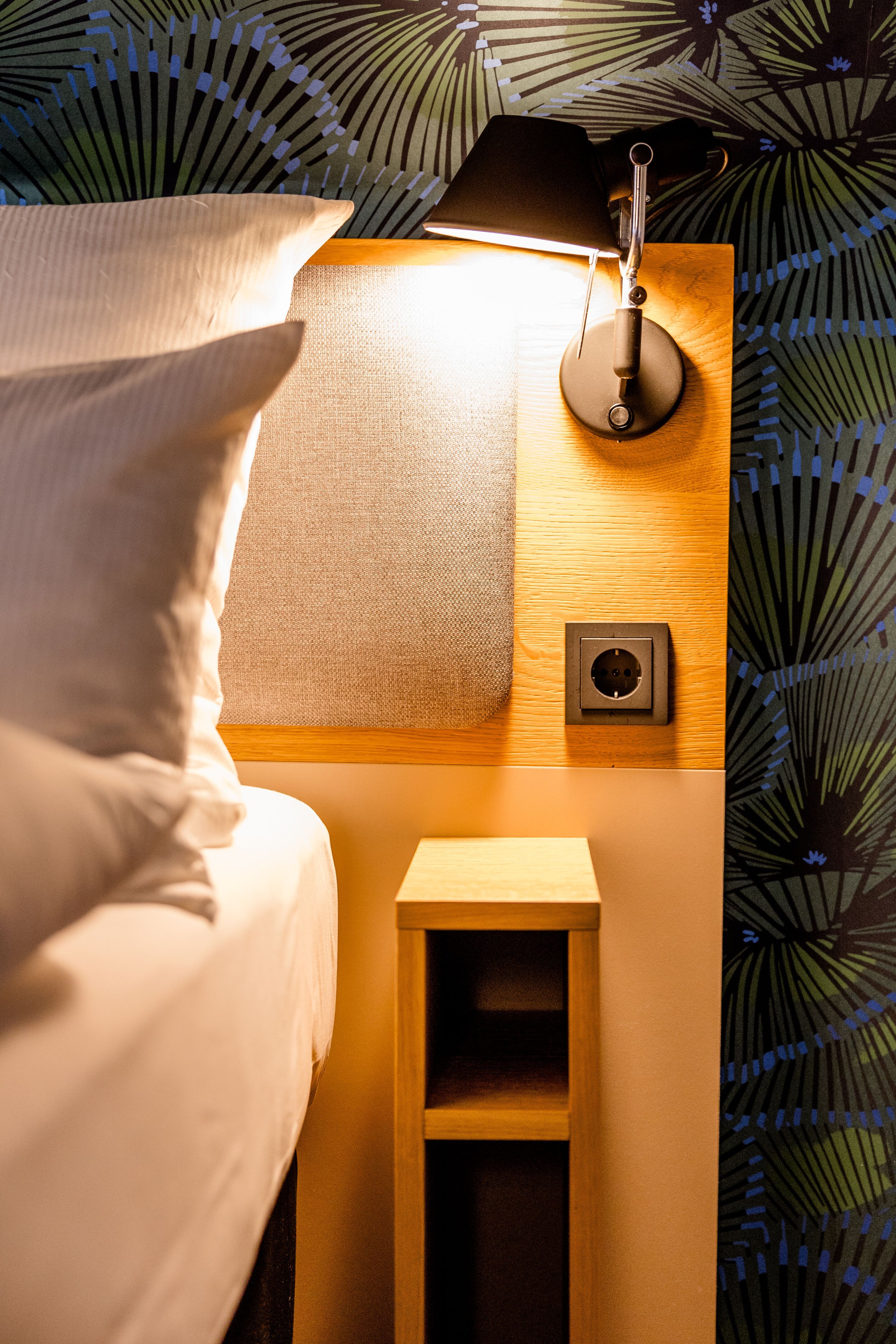 Hotel Frankfurt Hauptbahnhof Motel One beleuchtetes Kopfteil eines Hotelbetts mit weichen Kissen, Wandlicht und praktischem Nachttisch, umgeben von gemusterter Tapete.