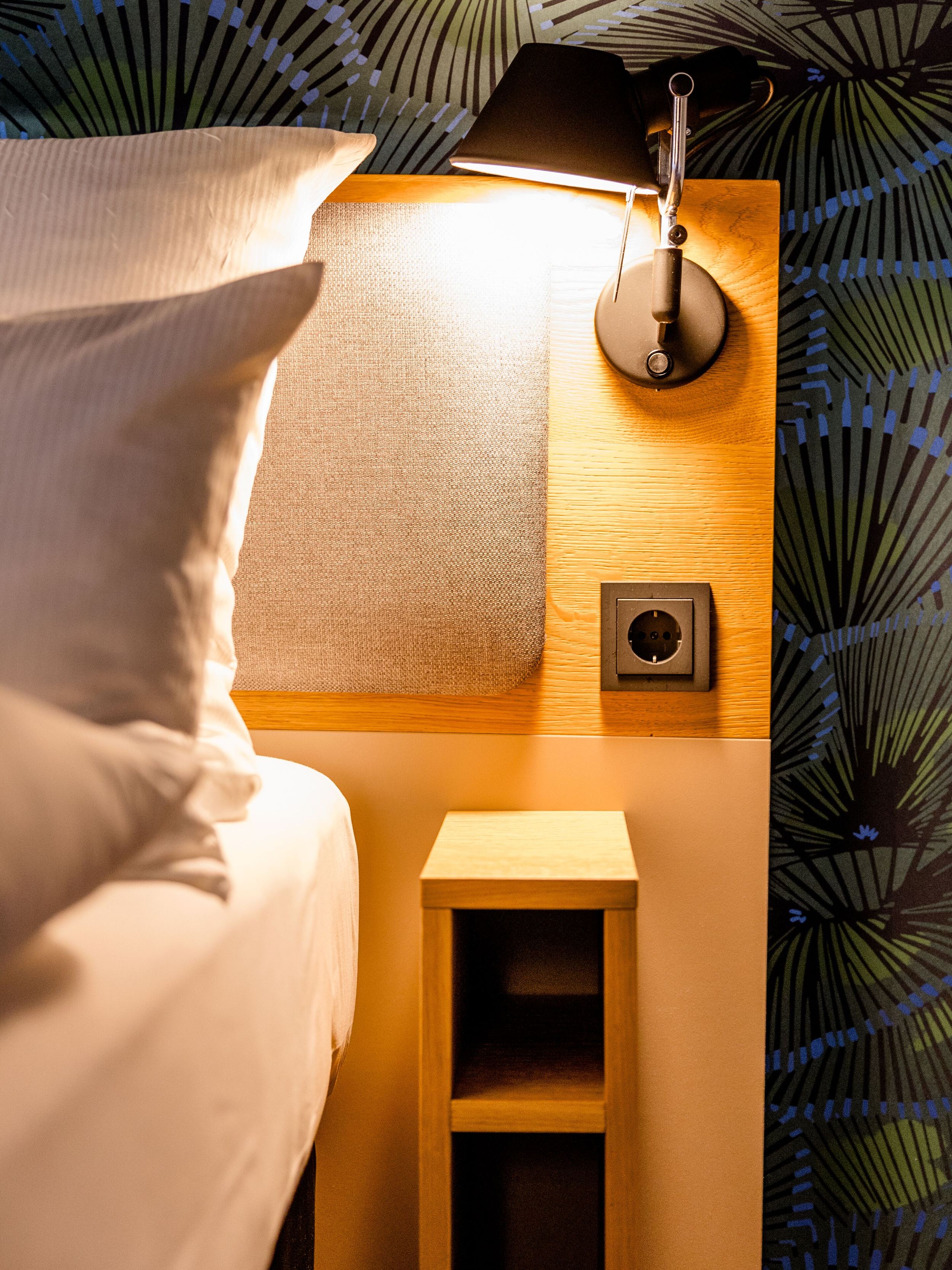 Hotel Frankfurt Hauptbahnhof Motel One beleuchtetes Kopfteil eines Hotelbetts mit weichen Kissen, Wandlicht und praktischem Nachttisch, umgeben von gemusterter Tapete.