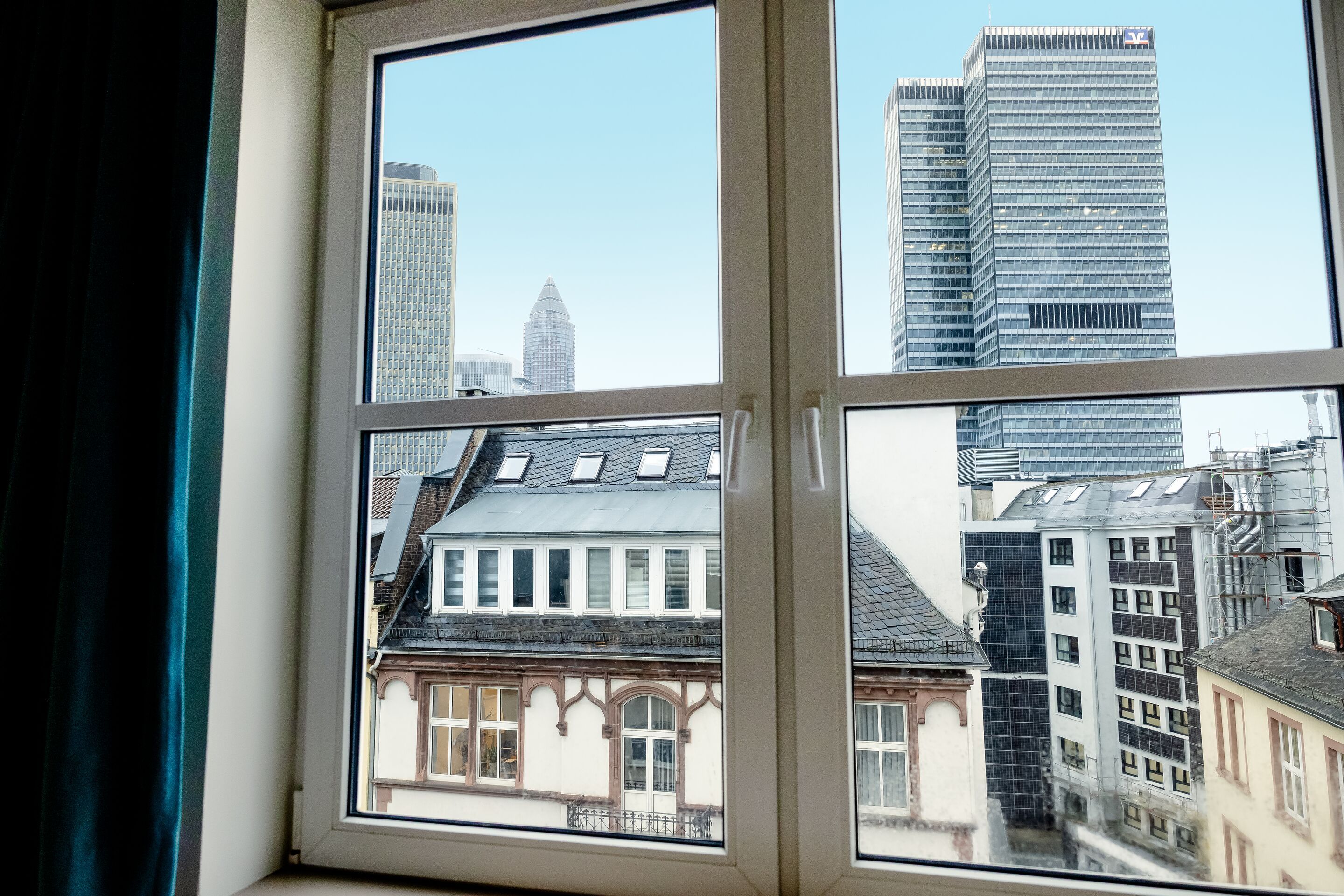 Hotel Frankfurt Hauptbahnhof Motel One Blick aus dem Hotelzimmerfenster auf die Frankfurter Skyline mit modernen Hochhäusern und historischen Gebäuden, bei klarem Himmel.
