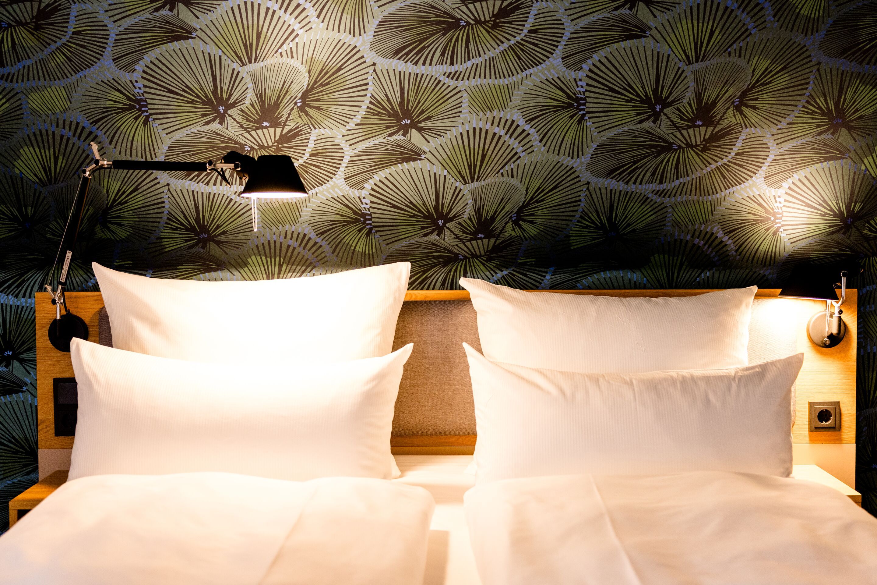 Hotel Frankfurt Hauptbahnhof Motel One beleuchtetes Doppelbett mit weißen Kissen und Bettwäsche vor einer stilvollen, dunkelgrünen Wand mit floralem Muster, zwei Leselampen sorgen für gemütliche Atmosphäre.