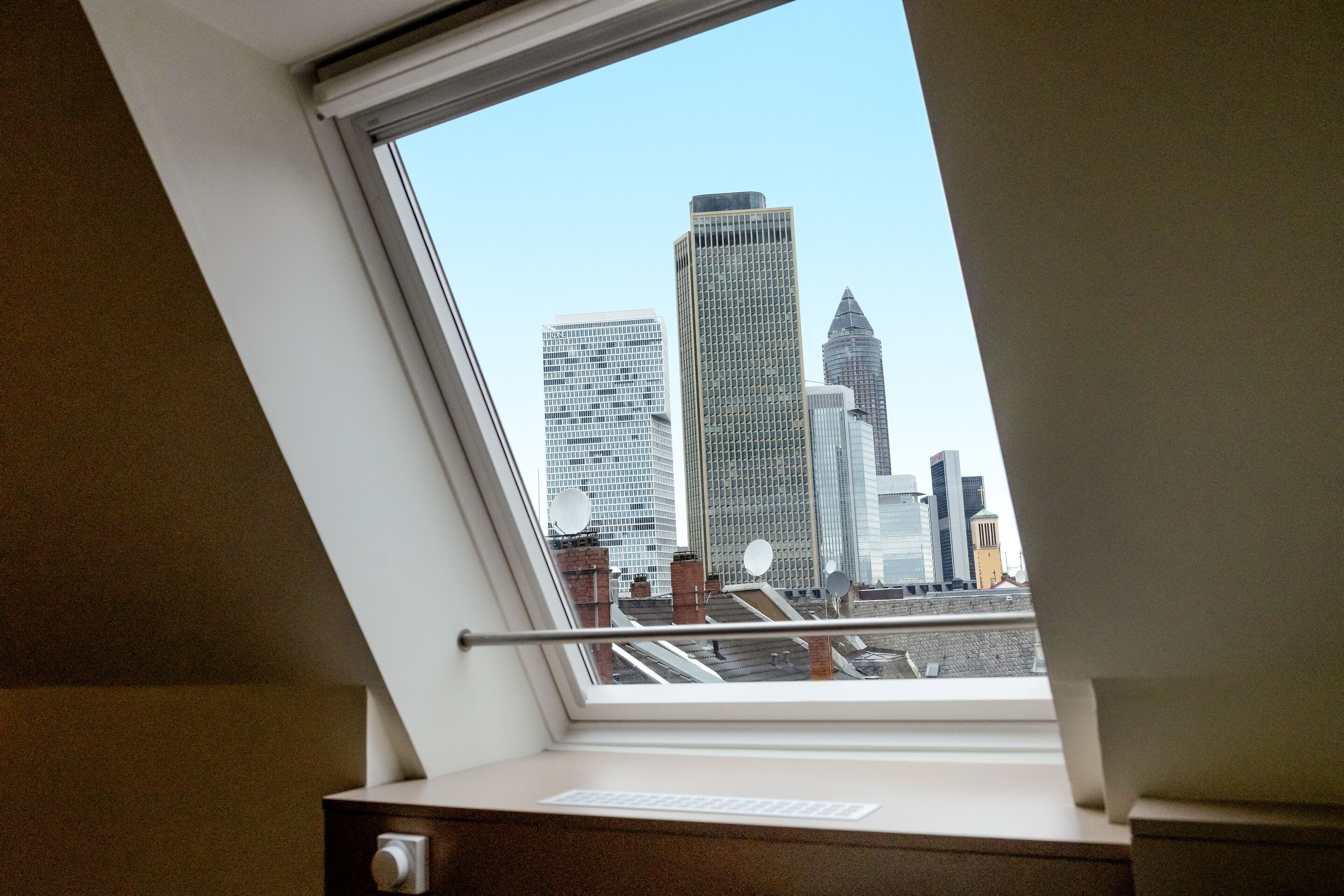 Hotel Frankfurt Hauptbahnhof Motel One Zimmer mit Blick aus einem Dachfenster auf die Frankfurter Skyline mit mehreren Hochhäusern bei klarem Himmel.