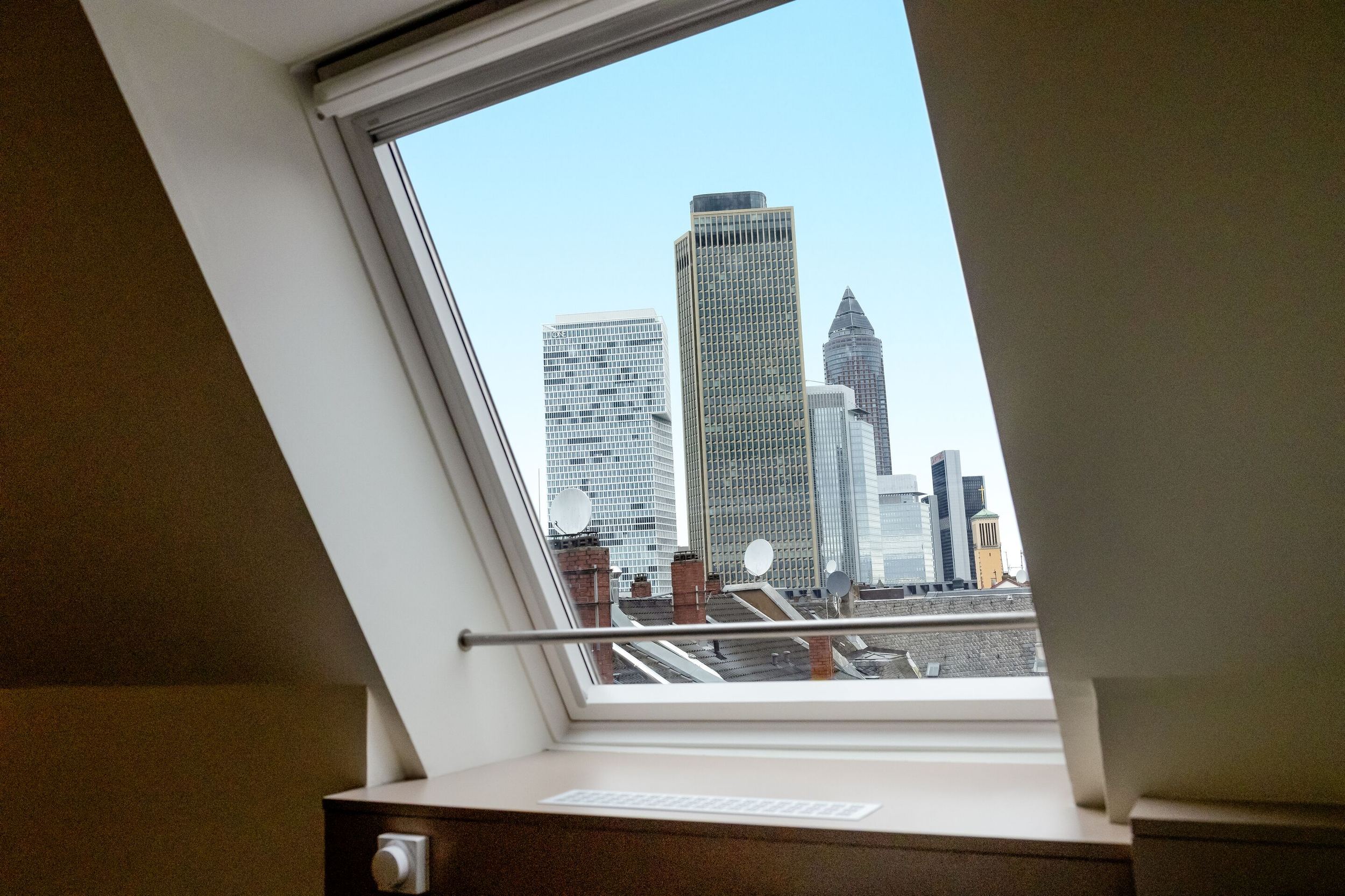 Hotel Frankfurt Hauptbahnhof Motel One Zimmer mit Blick aus einem Dachfenster auf die Frankfurter Skyline mit mehreren Hochhäusern bei klarem Himmel.