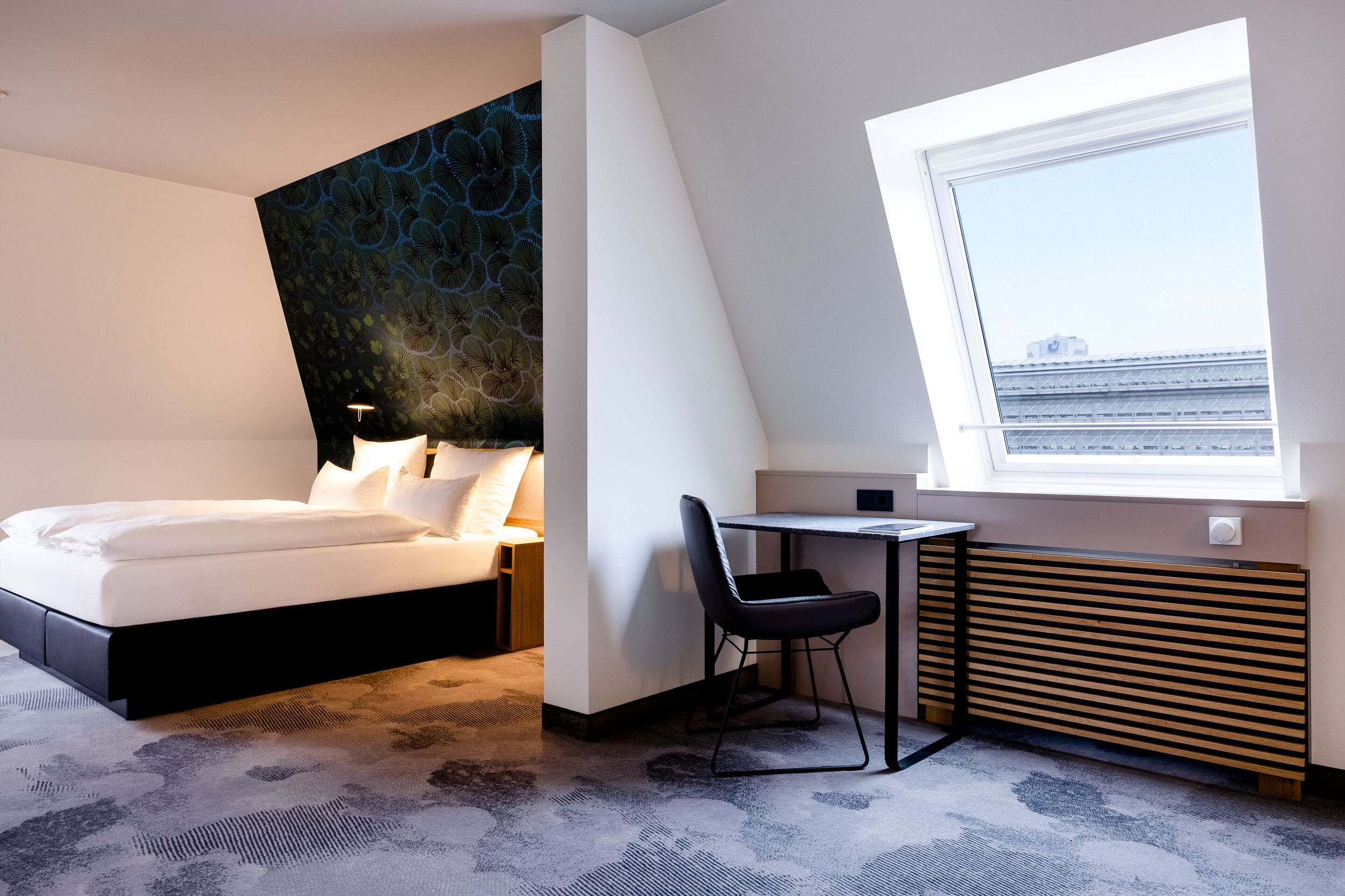 Hotel Frankfurt Hauptbahnhof Motel One Hotelzimmer mit großem, bequemem Doppelbett, stilvoller dunkler Akzentwand, Schreibtisch am Fenster mit Tageslicht und gemütlicher Atmosphäre für erholsamen Aufenthalt und produktives Arbeiten.