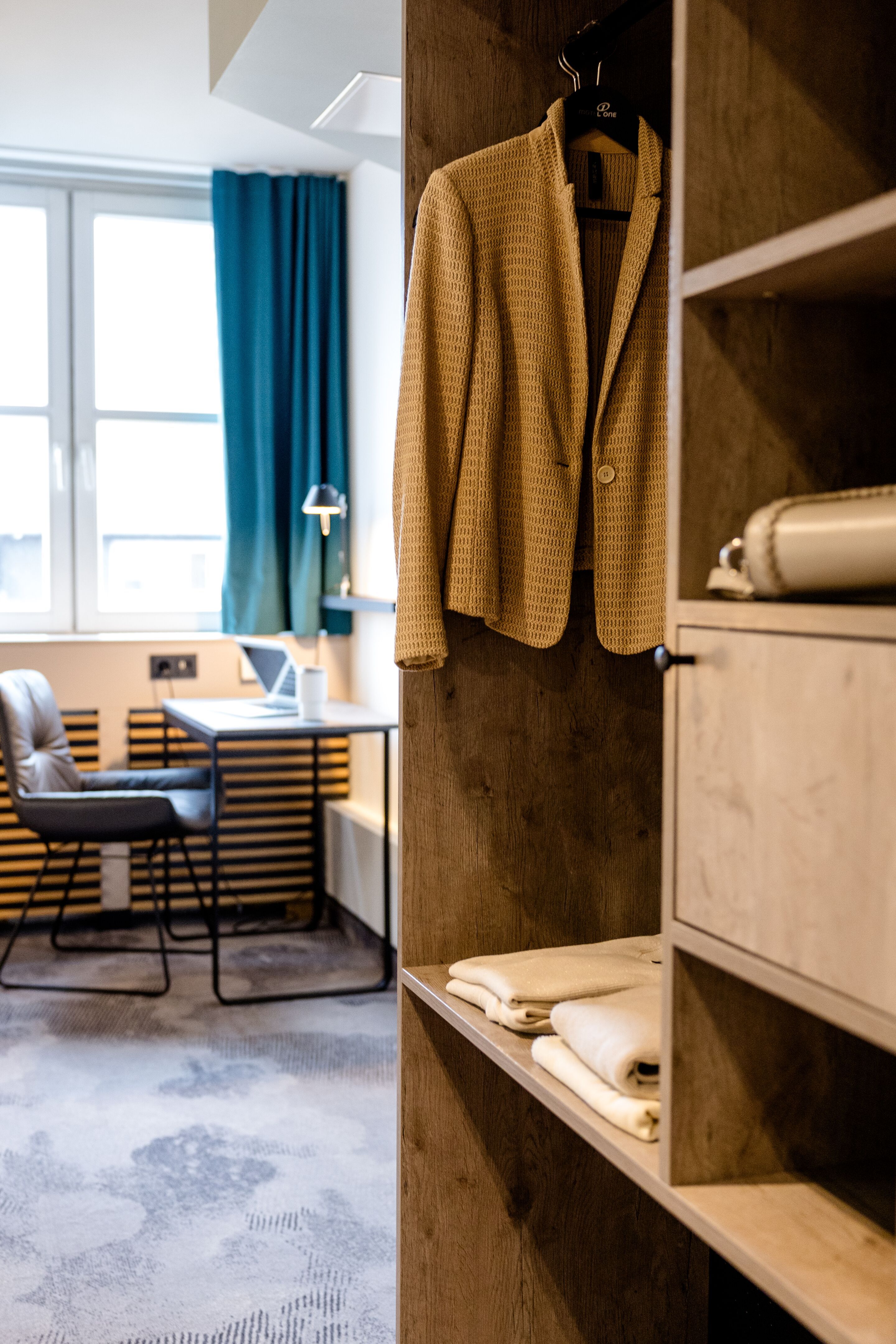 Designhotel Frankfurt Hauptbahnhof Motel One offener Kleiderschrank mit Jacke und Faltwäsche, im Hintergrund ein helles Zimmer mit Schreibtisch, Stuhl und großem Fenster für komfortables Arbeiten und Entspannen.