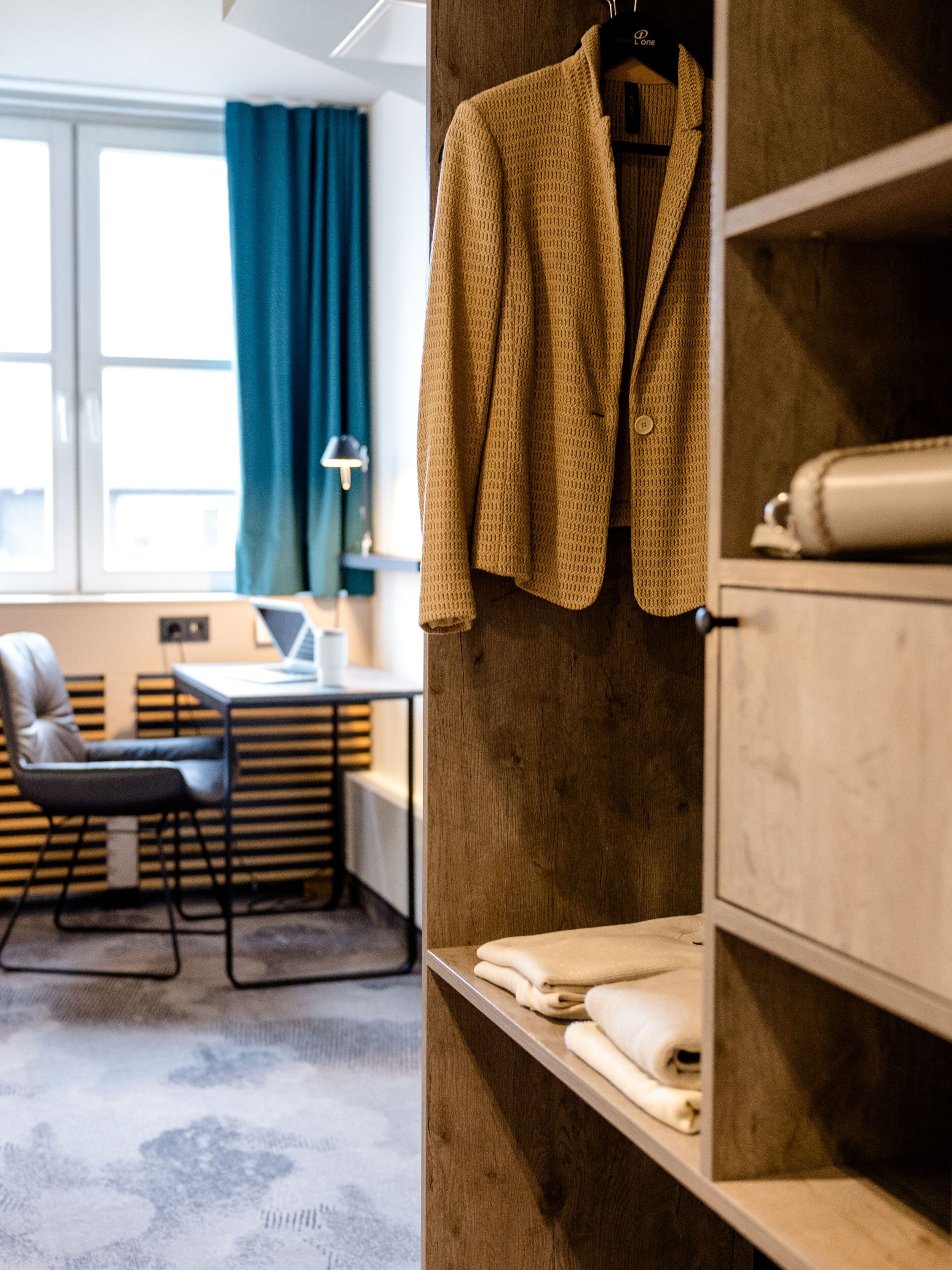 Designhotel Frankfurt Hauptbahnhof Motel One offener Kleiderschrank mit Jacke und Faltwäsche, im Hintergrund ein helles Zimmer mit Schreibtisch, Stuhl und großem Fenster für komfortables Arbeiten und Entspannen.