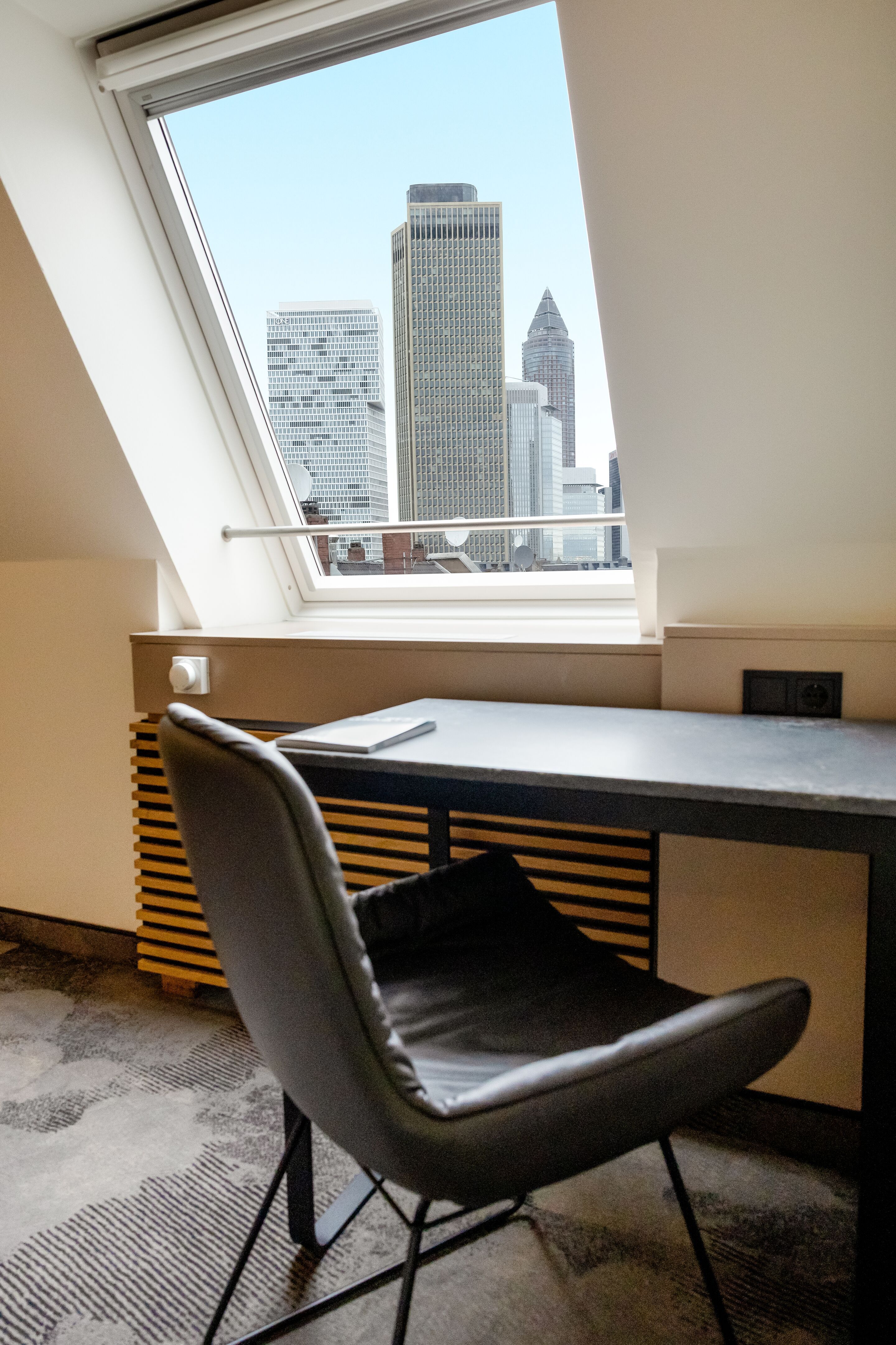 Hotel Frankfurt Hauptbahnhof Motel One Zimmer mit Arbeitsplatz mit schwarzem Stuhl und Schreibtisch vor einem großen Fenster, das eine klare Sicht auf die Hochhäuser von Frankfurt bietet.