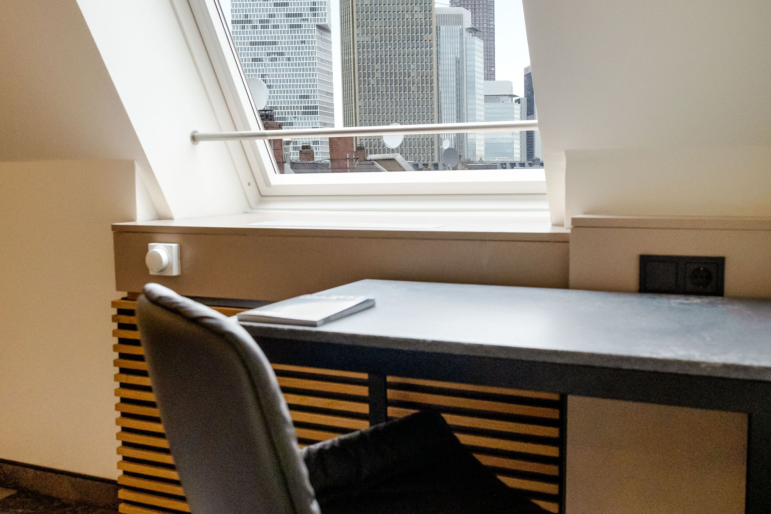 Hotel Frankfurt Hauptbahnhof Motel One Zimmer mit Arbeitsplatz mit schwarzem Stuhl und Schreibtisch vor einem großen Fenster, das eine klare Sicht auf die Hochhäuser von Frankfurt bietet.