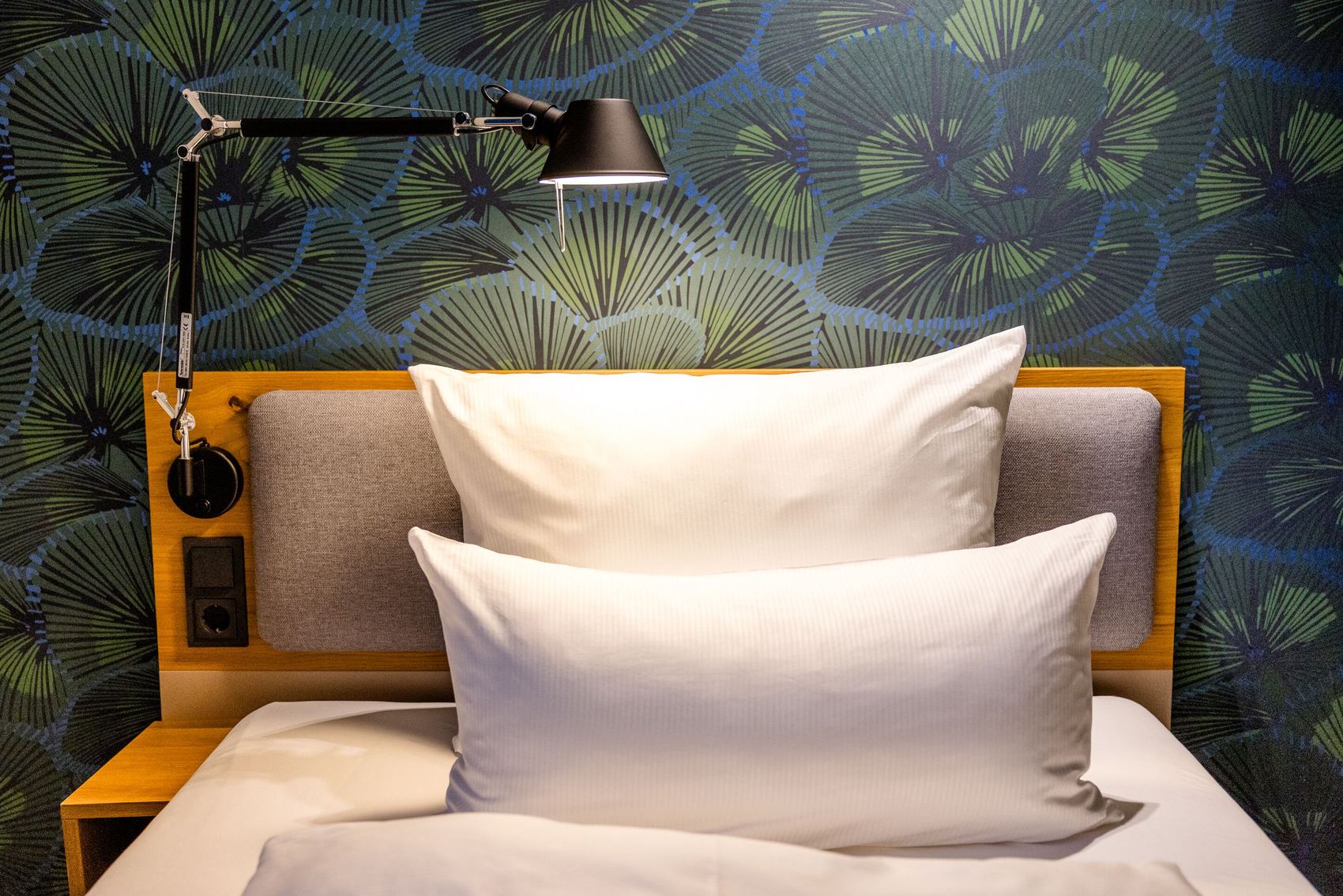 Hotel Motel One Frankfurt-Hauptbahnhof Gemütliches Hotelbett mit zwei weißen Kissen, gepolstertem Kopfteil und einer verstellbaren Leselampe vor einer stilvollen, dunkelgrünen Wand mit Blattmuster.