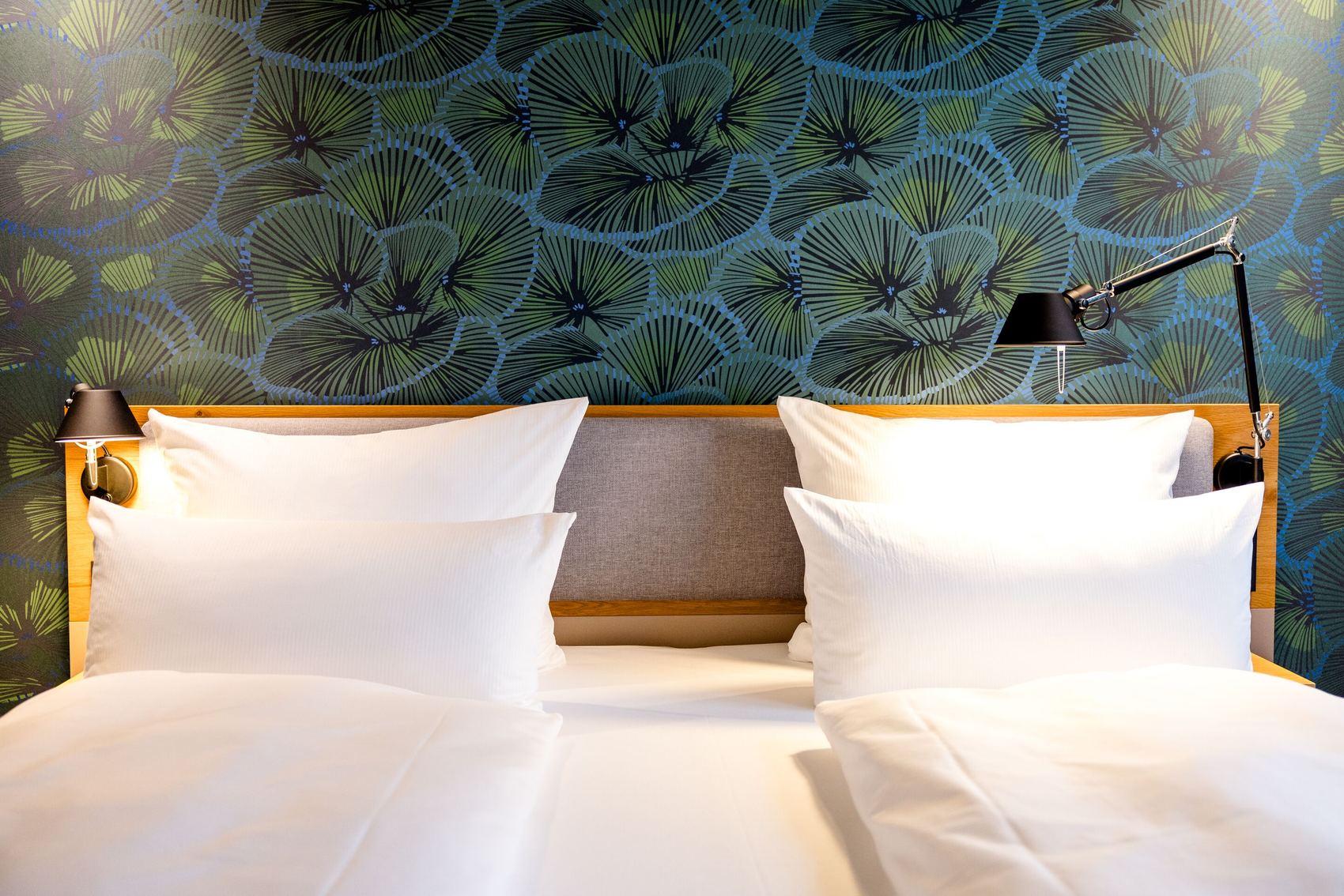 Hotel Motel One Frankfurt-Hauptbahnhof Modernes Hotelbett mit weißen Bettwaren vor einer Wand mit stilisiertem Blattmuster. An beiden Seiten Leselampen für eine gemütliche Atmosphäre.