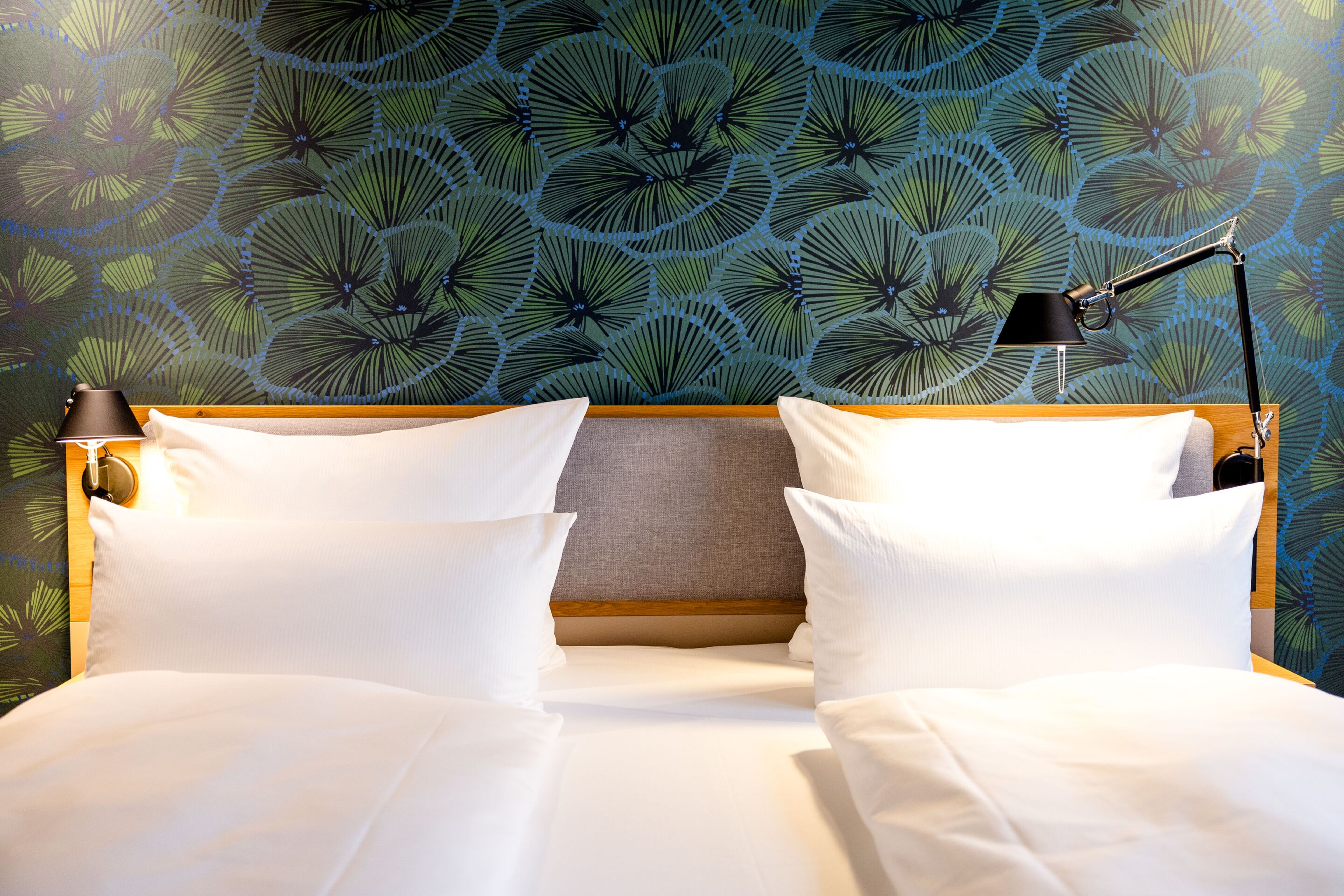 Hotel Motel One Frankfurt-Hauptbahnhof Modernes Hotelbett mit weißen Bettwaren vor einer Wand mit stilisiertem Blattmuster. An beiden Seiten Leselampen für eine gemütliche Atmosphäre. Hotel Motel One Frankfurt-Hauptbahnhof Modernes Hotelbett mit weißen Bettwaren vor einer Wand mit stilisiertem Blattmuster. An beiden Seiten Leselampen für eine gemütliche Atmosphäre.