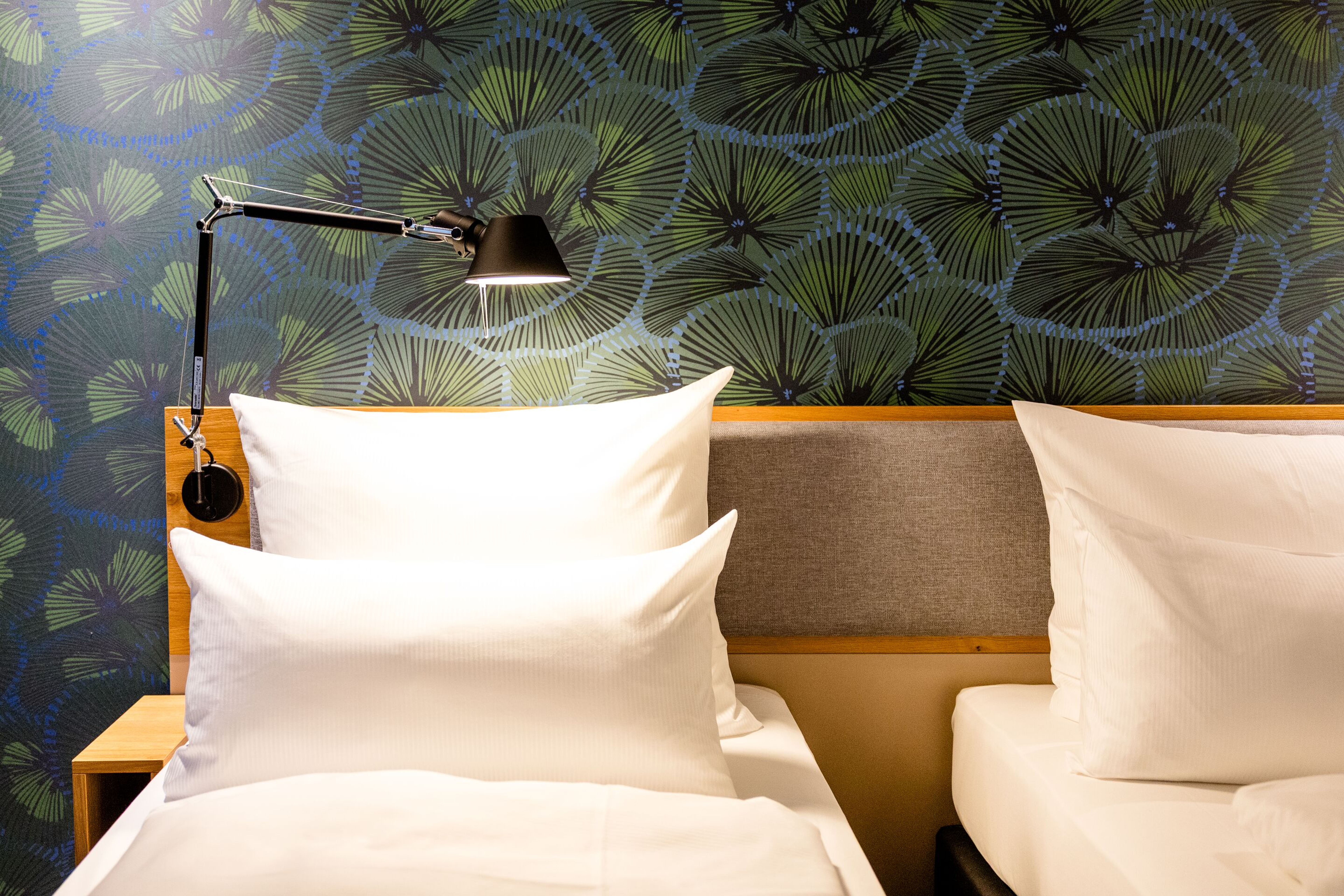 Hotel Motel One Frankfurt-Hauptbahnhof Doppelbett mit weißen Kissen und Bettwäsche vor einer Wand mit stilisiertem grün-blauem Blattmuster, daneben eine moderne Leselampe, die eine gemütliche Schlafraum-Atmosphäre schafft. Hotel Motel One Frankfurt-Hauptbahnhof Doppelbett mit weißen Kissen und Bettwäsche vor einer Wand mit stilisiertem grün-blauem Blattmuster, daneben eine moderne Leselampe, die eine gemütliche Schlafraum-Atmosphäre schafft.
