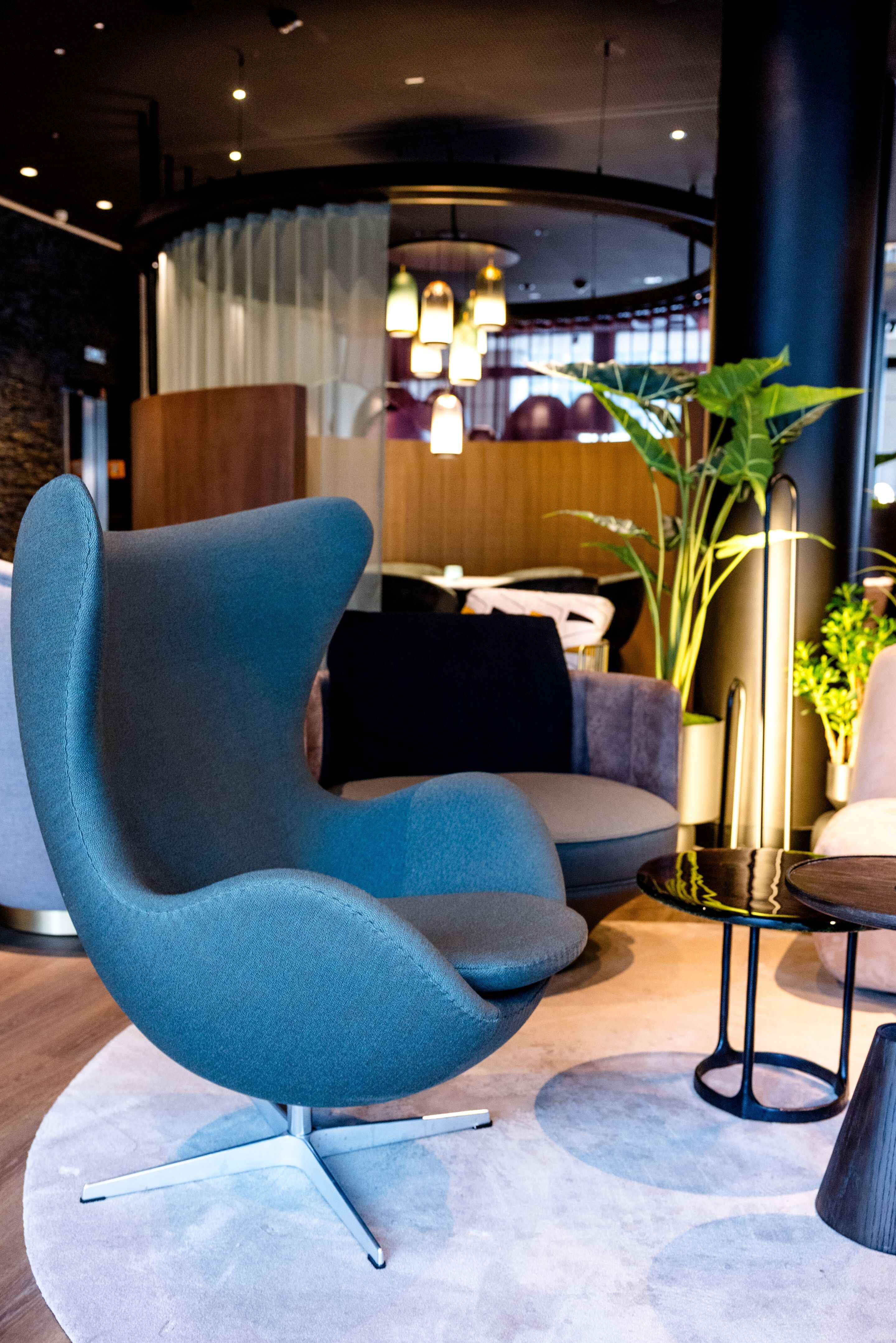 Hotel Frankfurt-Messe Motel One Lounge mit Egg Chair im Vordergrund, rundem Teppich, eleganten Beistelltischen sowie wohltuende Beleuchtung und grüne Pflanzen.