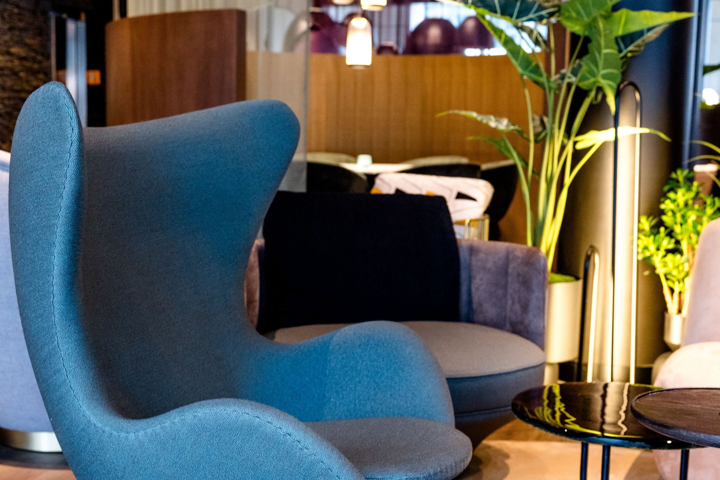 Hotel Frankfurt-Messe Motel One Lounge mit Egg Chair im Vordergrund, rundem Teppich, eleganten Beistelltischen sowie wohltuende Beleuchtung und grüne Pflanzen.