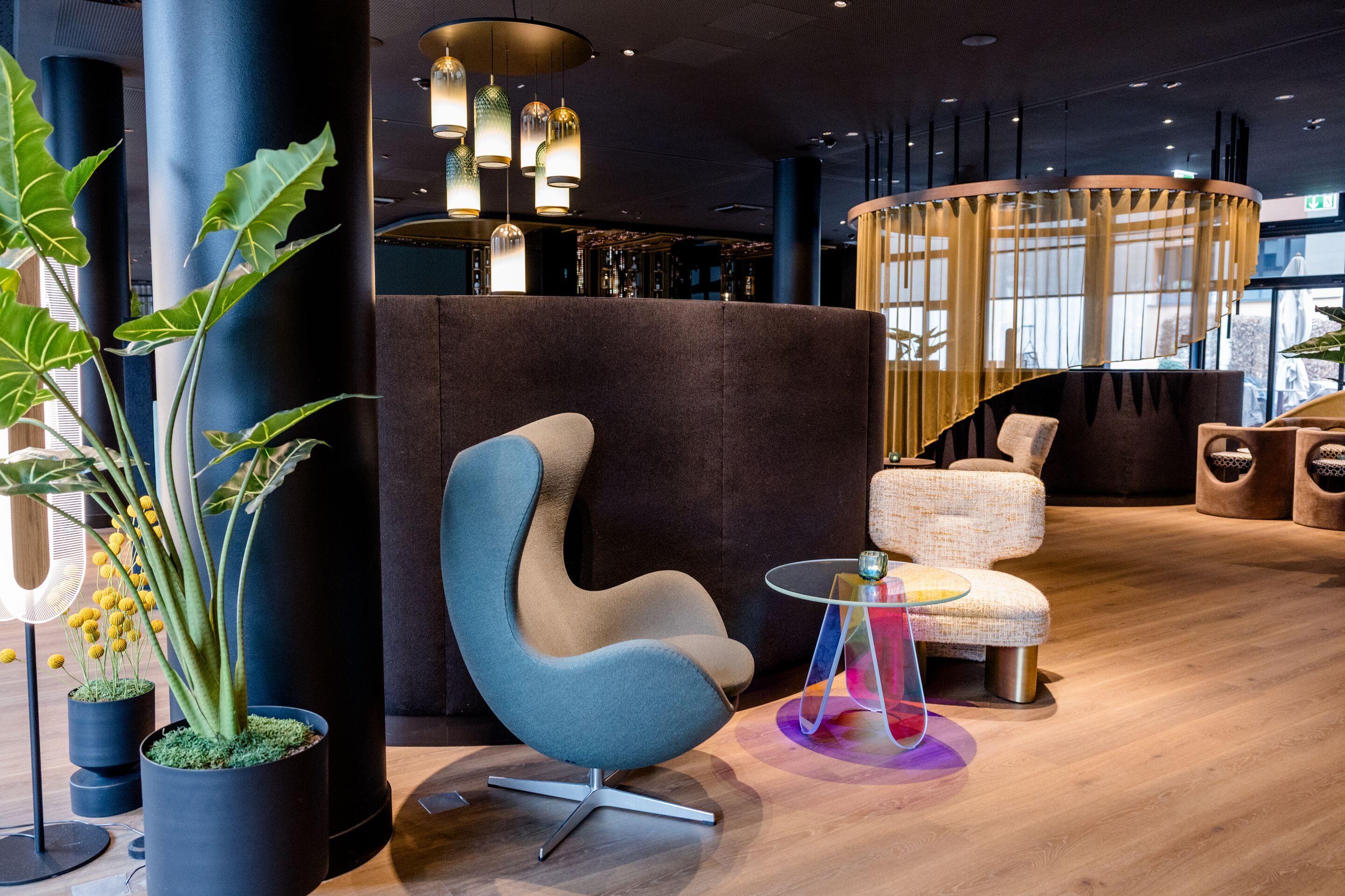 Hotel Frankfurt-Messe Motel One große Lounge mit mehreren Sesseln, kleinem Glastisch und warmem Holzboden, ergänzt durch dekorative Pflanzen und sanfte Beleuchtung.