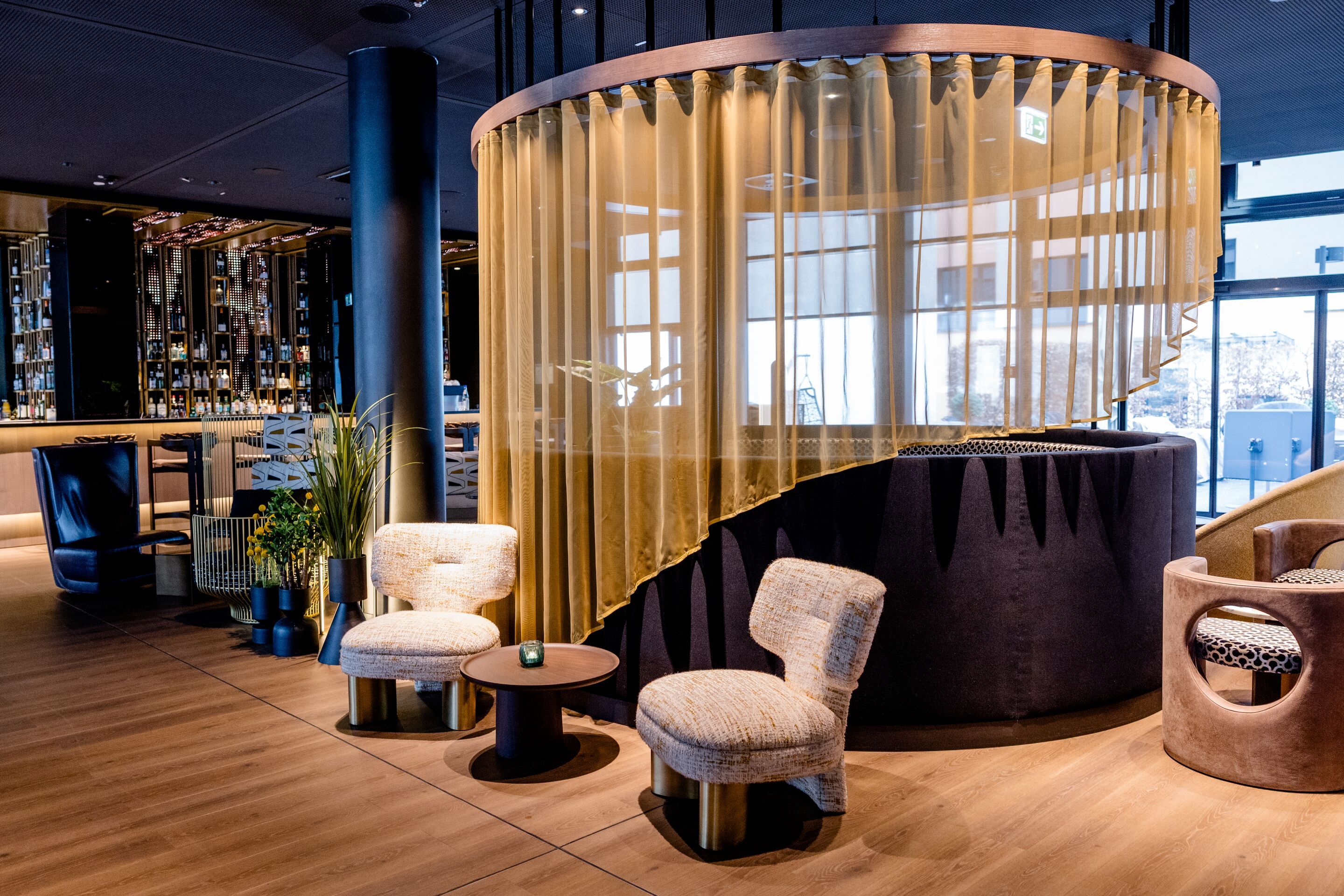 Hotel Frankfurt-Messe Motel One Lounge und Bar mit Sitzgruppe aus bequemen Sesseln, rundem Tisch und halbtransparentem Vorhang. Warme Beleuchtung und elegante Holz- und Stoffelemente.