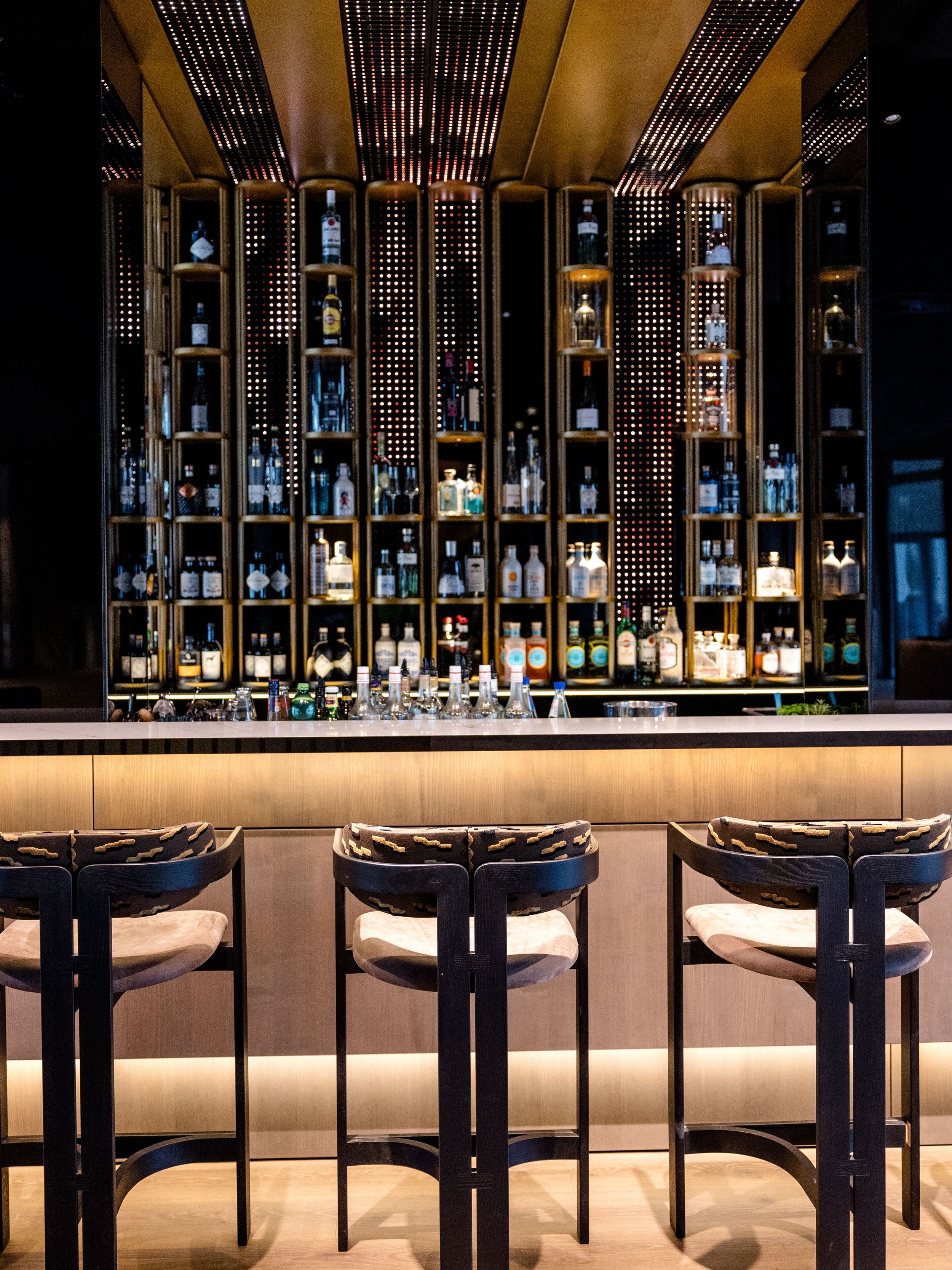 Hotel Frankfurt-Messe Motel One Bar mit Barhockern, beleuchtetem Tresen und Wandregalen voller hochwertiger Spirituosen, die eine stilvolle Atmosphäre für entspannte Abende schaffen.