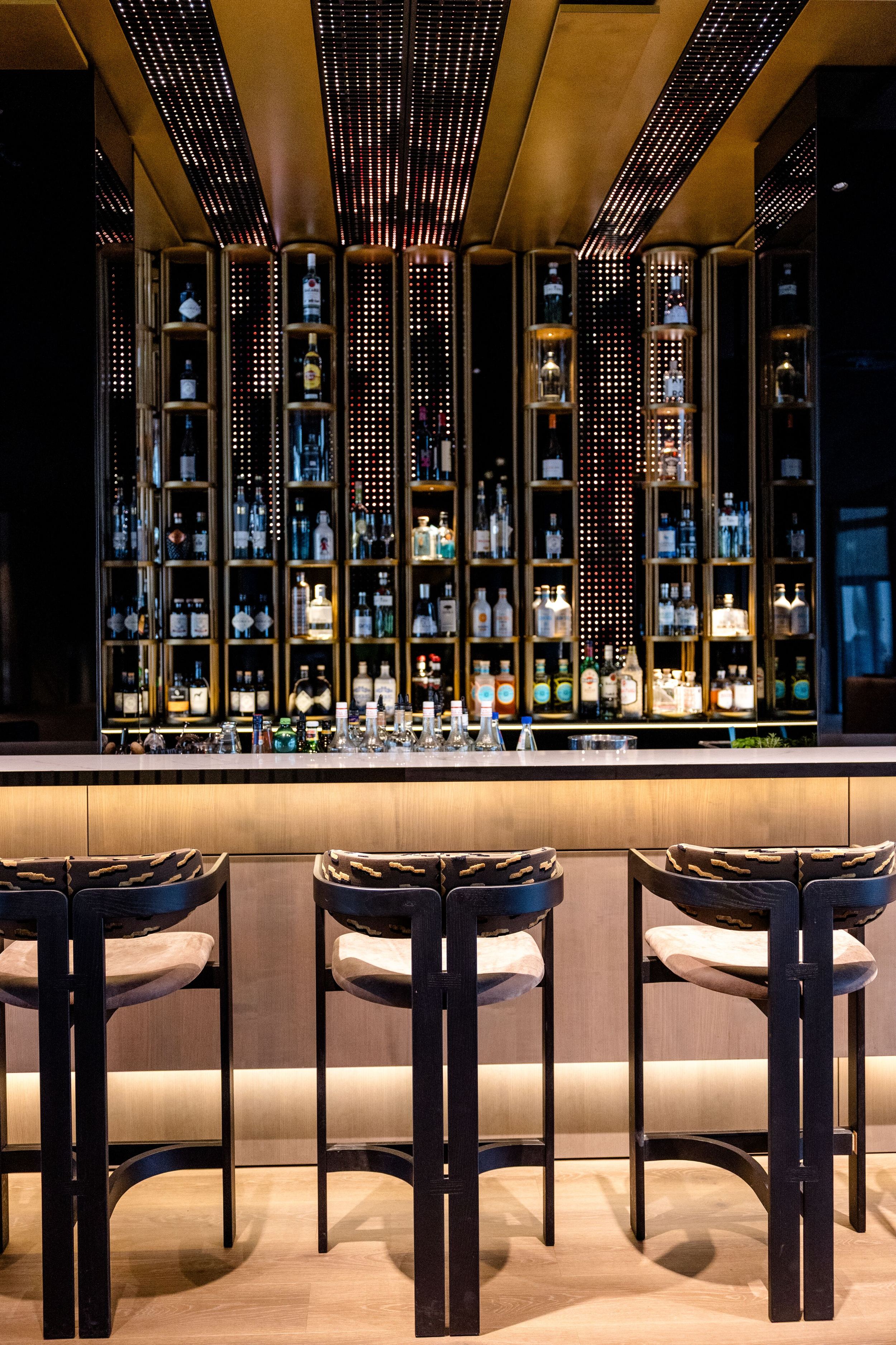 Hotel Frankfurt-Messe Motel One Bar mit Barhockern, beleuchtetem Tresen und Wandregalen voller hochwertiger Spirituosen, die eine stilvolle Atmosphäre für entspannte Abende schaffen.