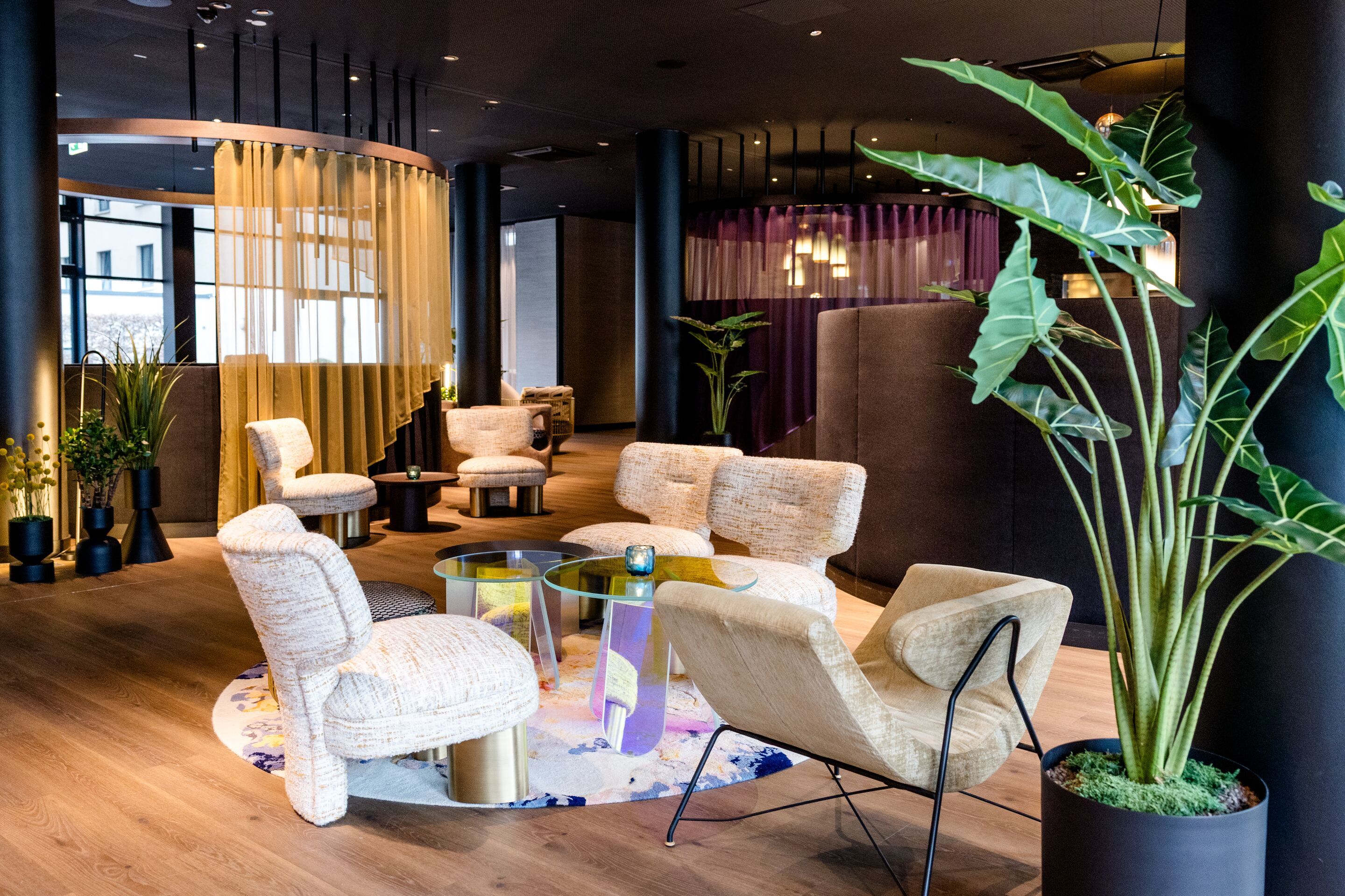 Designhotel Frankfurt-Messe Motel One Lounge mit bequemen Sesseln, runden Glastischen und dekorativen Pflanzen, der einladende Atmosphäre für Entspannung oder Gespräche bietet.