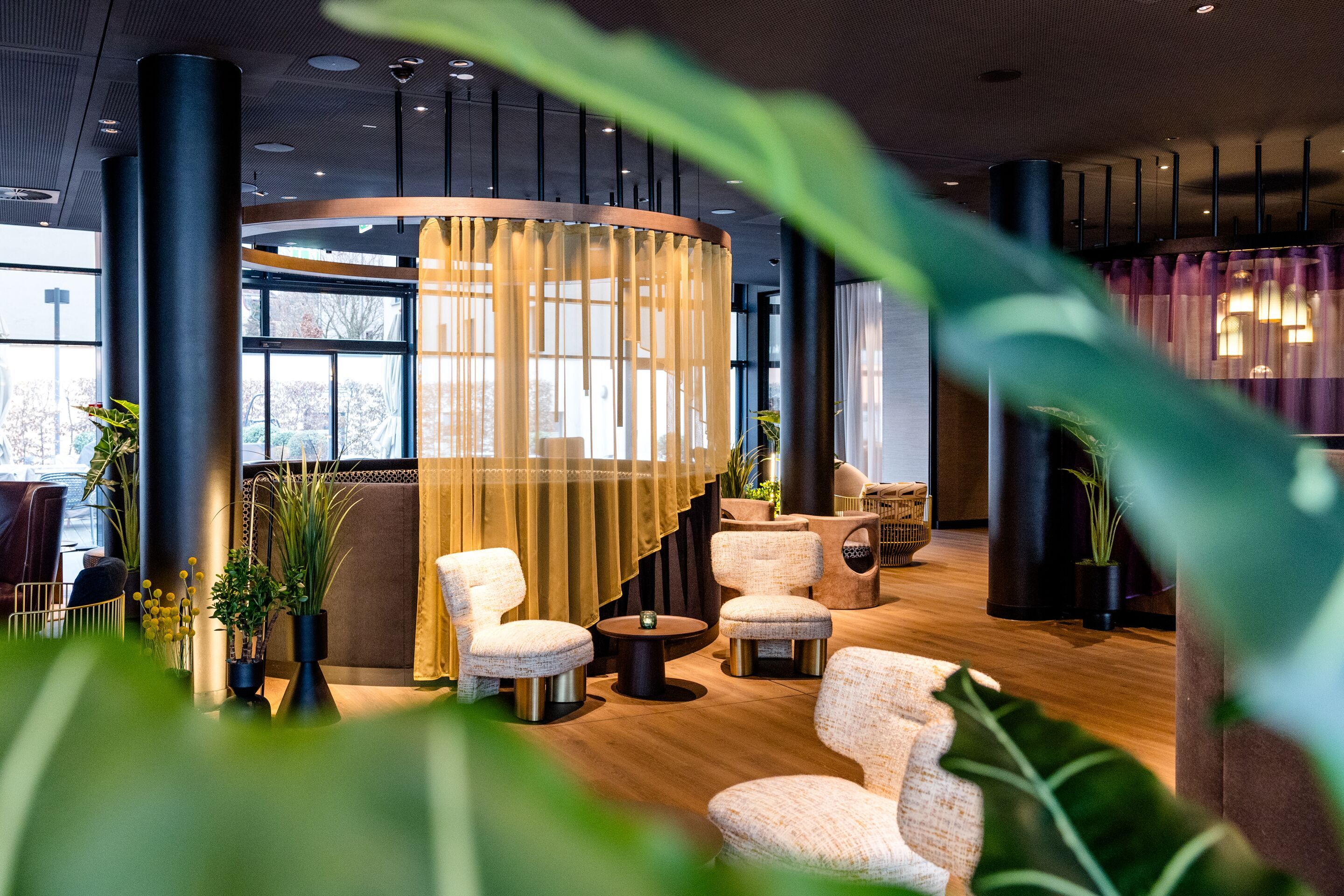 Hotel Frankfurt-Messe Motel One Lounge mit gemütlichen Sesseln, runden Tischen und dekorativen Pflanzen, umgeben von halbtransparenten Vorhängen, die für eine angenehme, entspannte Atmosphäre sorgen. Große Fenster bieten Tageslicht und Ausblicke nach draußen.