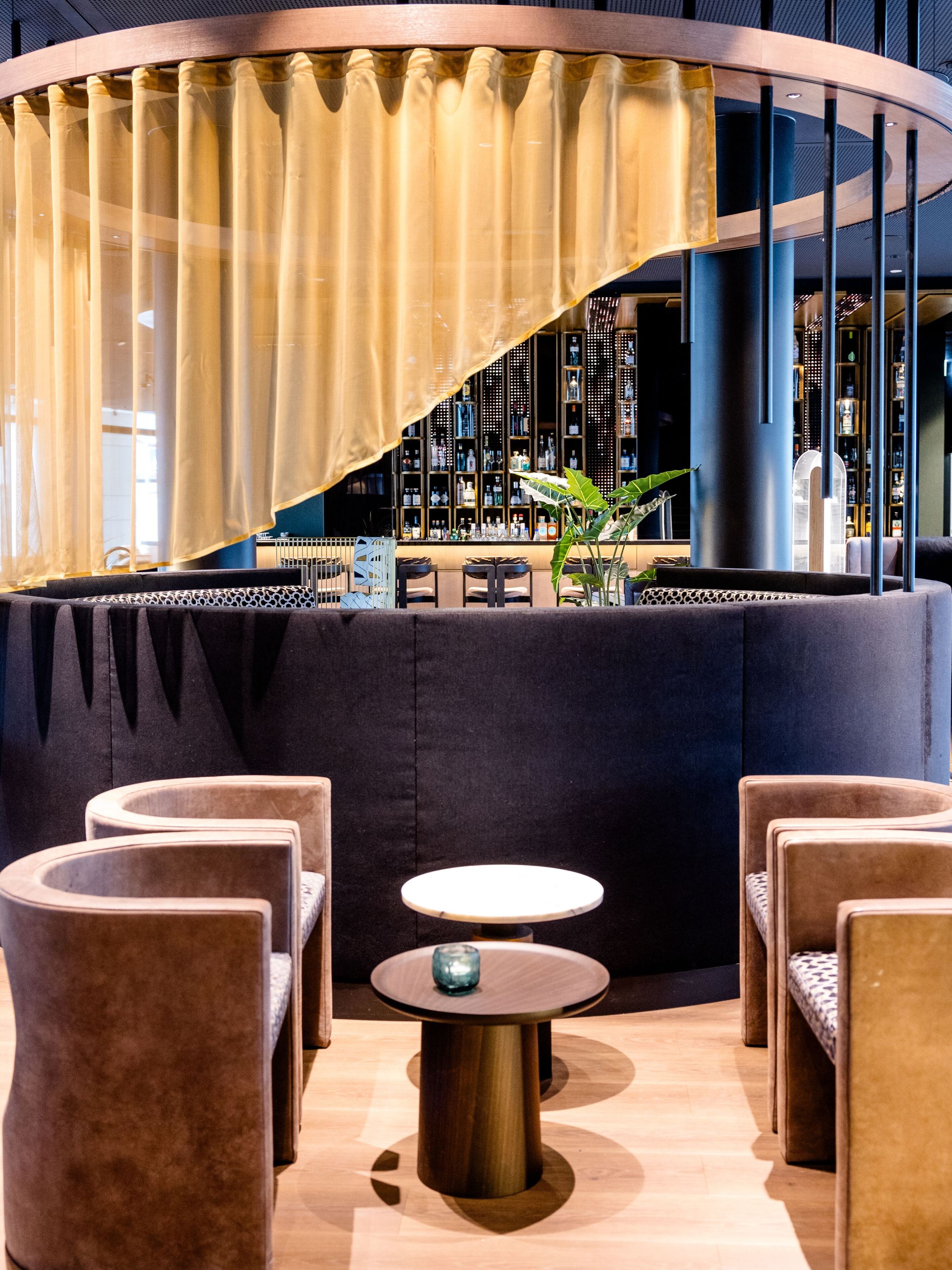 Hotel Frankfurt-Messe Motel One Loungbereich mit Sesseln und kleinen Tischen, abgetrennt durch einen halbtransparenten Vorhang, warmes, modernes Ambiente ideal zum Entspannen oder geselligen Beisammensein.