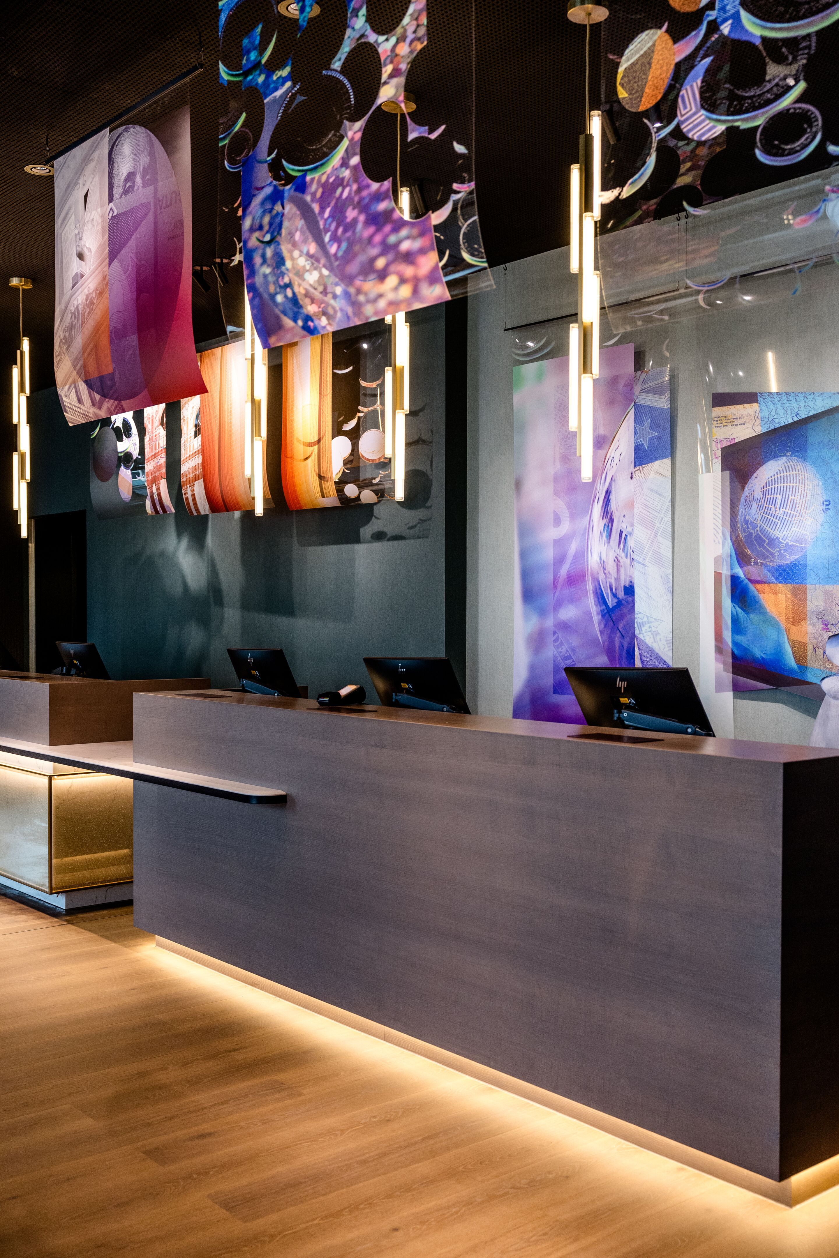 Designhotel Frankfurt-Messe Motel One Rezeption mit dunklem Tresen, indirekter Beleuchtung und farbenfroher, künstlerischer Wanddekoration.