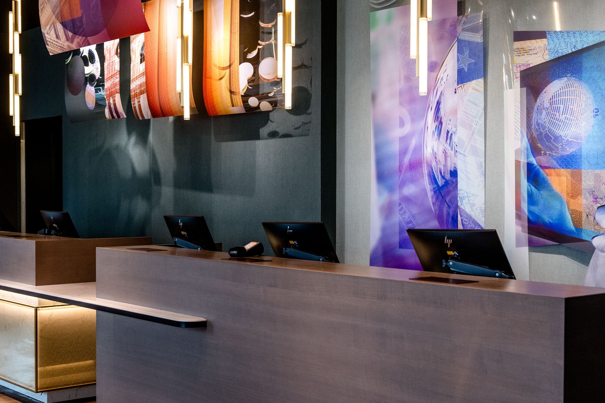 Designhotel Frankfurt-Messe Motel One Rezeption mit dunklem Tresen, indirekter Beleuchtung und farbenfroher, künstlerischer Wanddekoration.