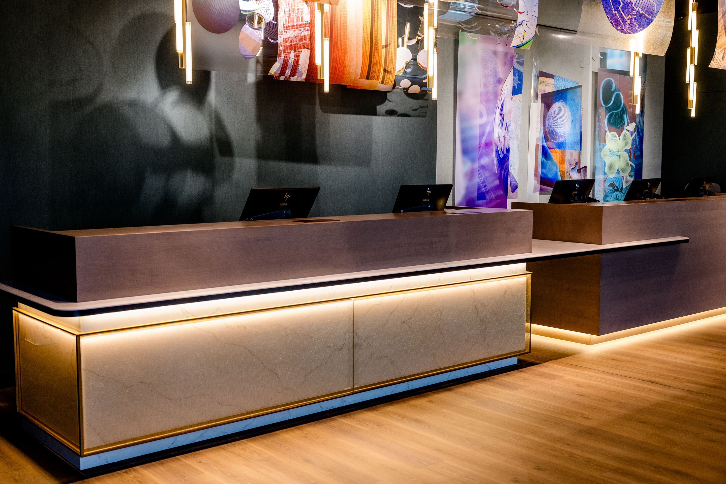 Hotel Frankfurt-Messe Motel One Empfangsbereich mit elegant beleuchtetem Tresen der Rezeption aus Holz und Marmor, warmem Lichtakzent, zeitgenössischer Kunst an den Wänden und einer einladenden Atmosphäre für Gäste.