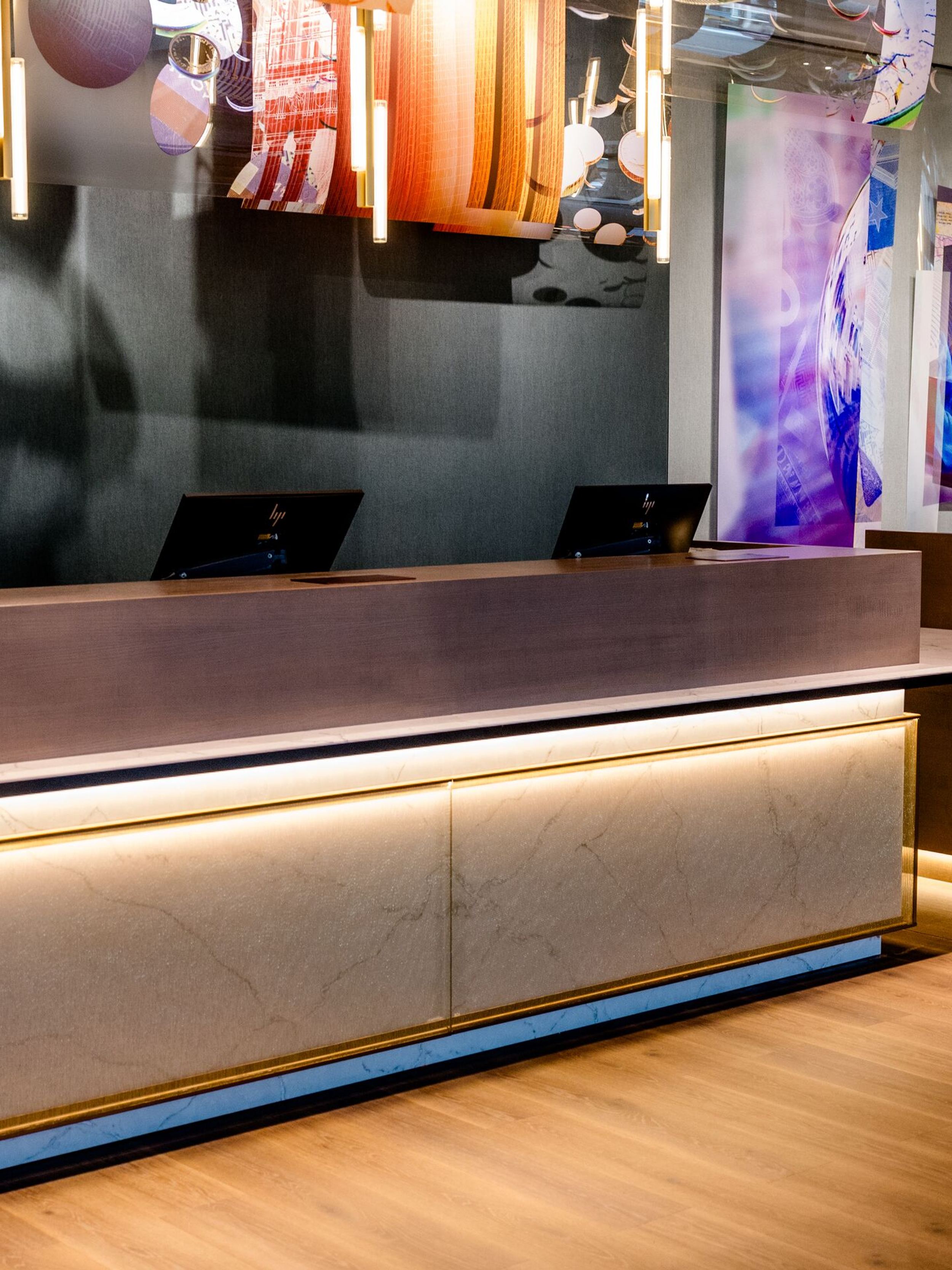 Hotel Frankfurt-Messe Motel One Empfangsbereich mit elegant beleuchtetem Tresen der Rezeption aus Holz und Marmor, warmem Lichtakzent, zeitgenössischer Kunst an den Wänden und einer einladenden Atmosphäre für Gäste.