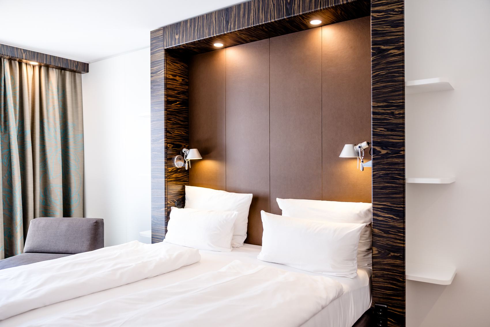 Modernes Hotelzimmer mit Doppelbett, hellem Bettzeug und gepolstertem Kopfteil, beleuchtet von integrierten Nachttischlampen, dazu ein gemütlicher Sessel und verdunkelnde Vorhänge am Fenster.