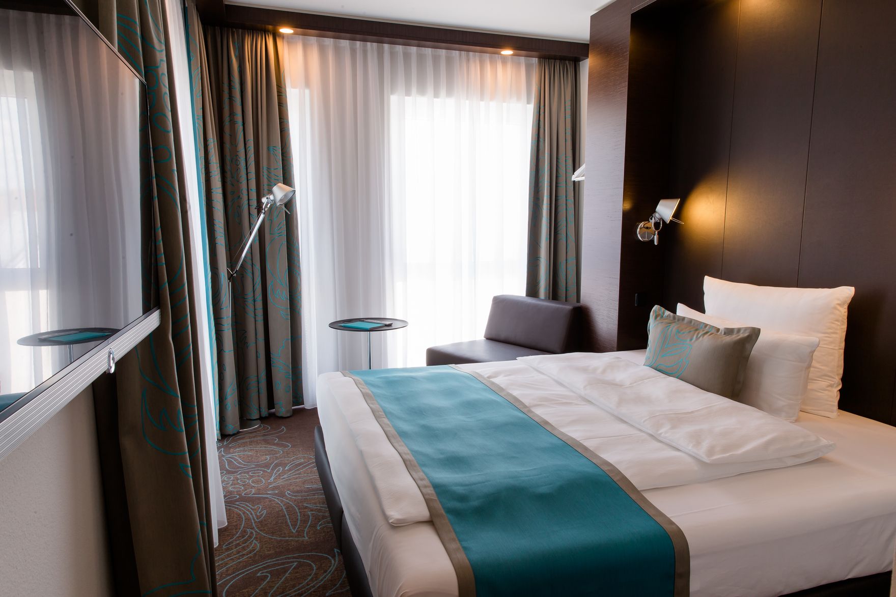 Hotel Frankfurt Messe Motel One seitlicher Blick in großes Doppelzimmer mit Bett, Sessel, Beistelltisch sowie Fernseher an der Wand