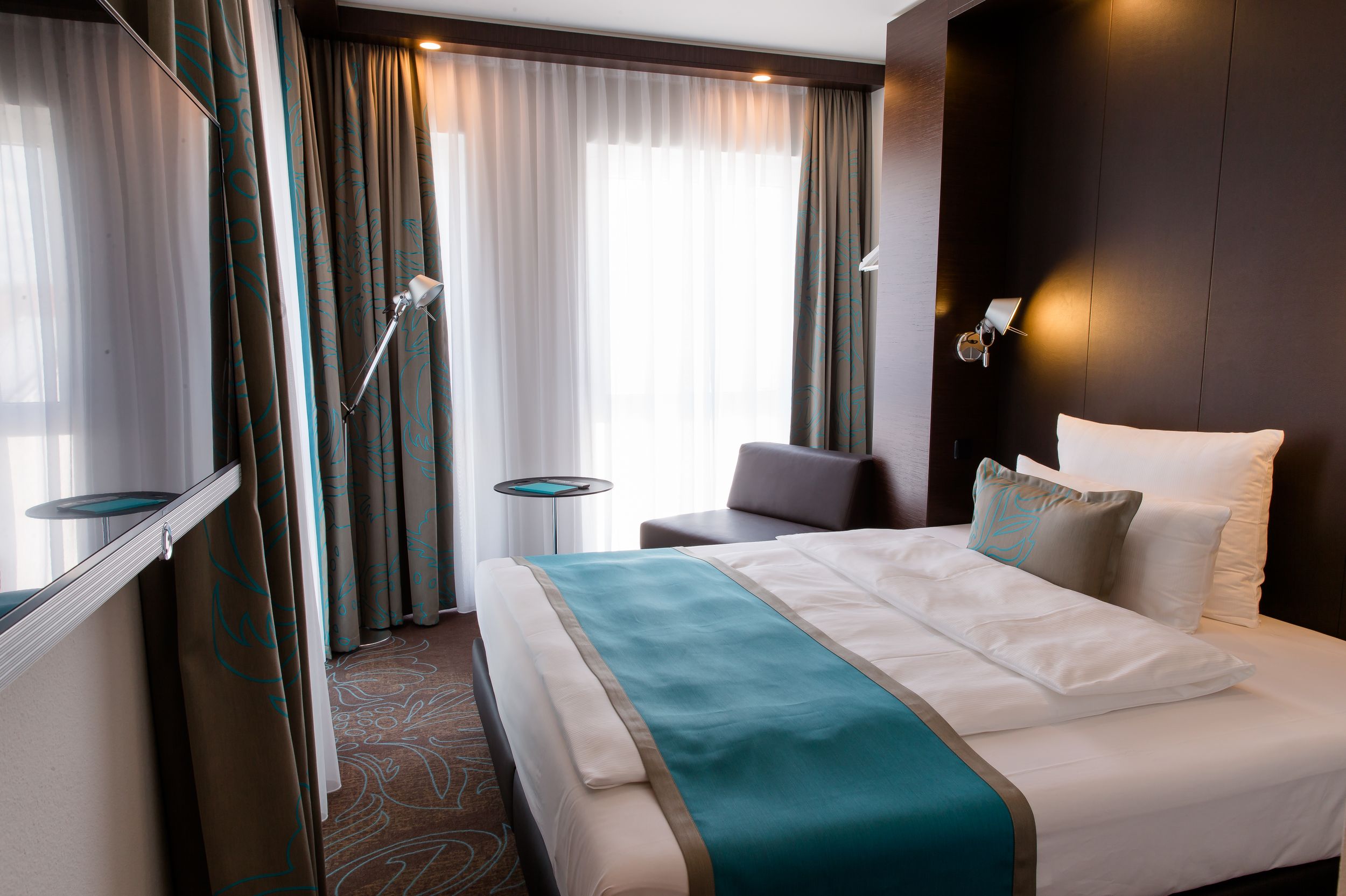 Hotel Frankfurt Messe Motel One seitlicher Blick in großes Doppelzimmer mit Bett, Sessel, Beistelltisch sowie Fernseher an der Wand