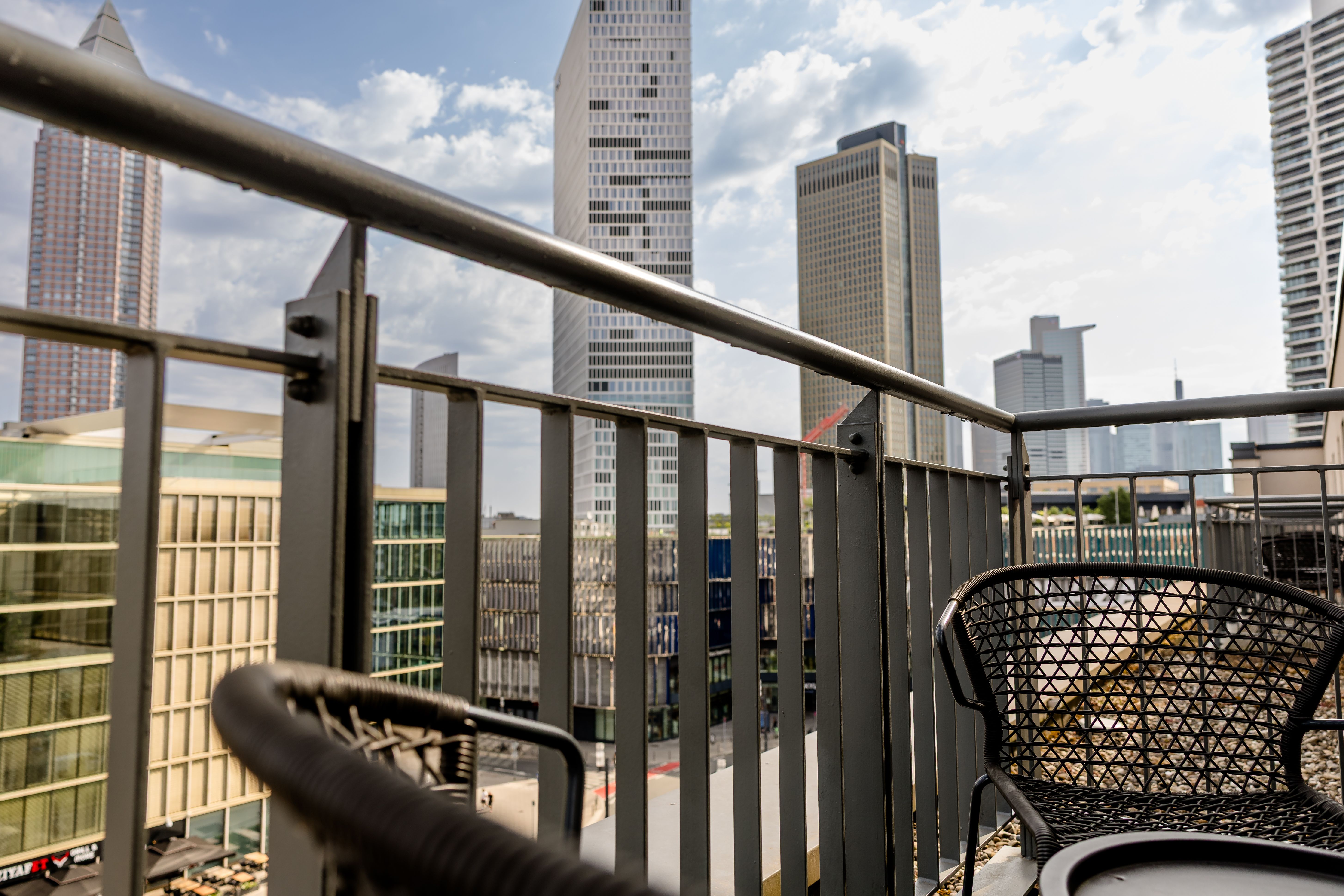 Balkon mit zwei Flechtsesseln und Blick auf moderne Hochhäuser einer Stadt, bei Tageslicht mit teils bewölktem Himmel.