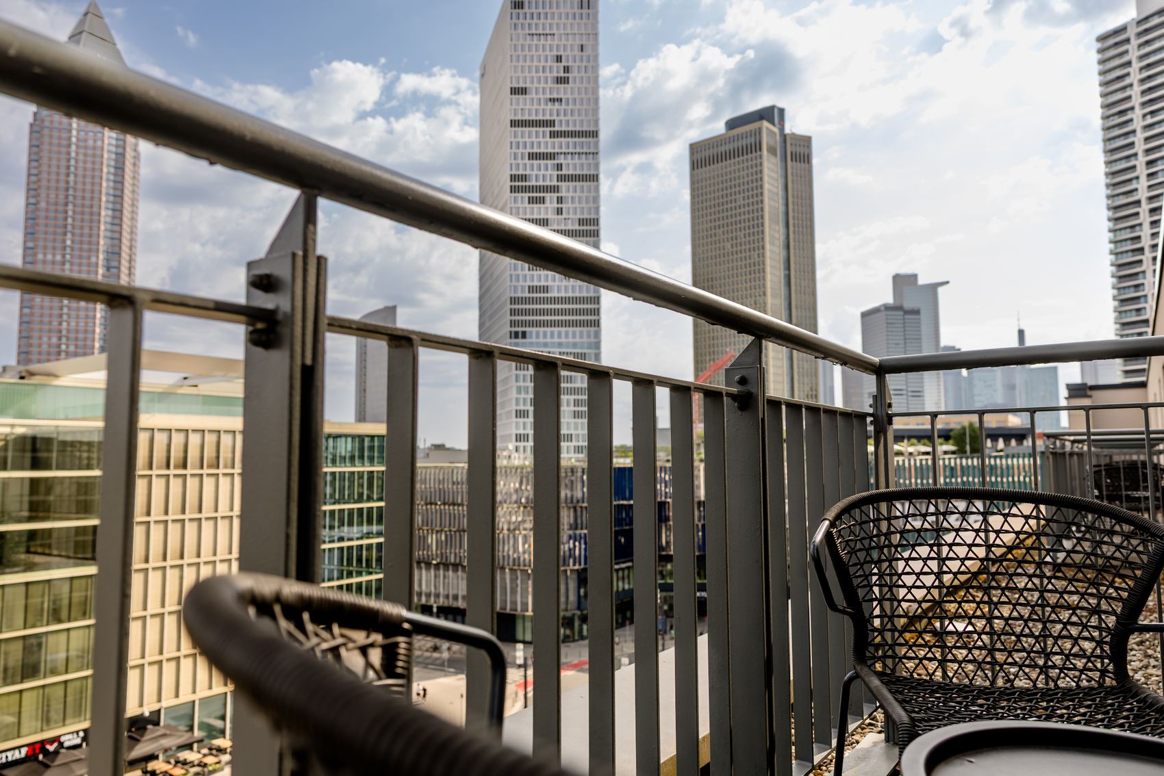 Balkon mit zwei Flechtsesseln und Blick auf moderne Hochhäuser einer Stadt, bei Tageslicht mit teils bewölktem Himmel.