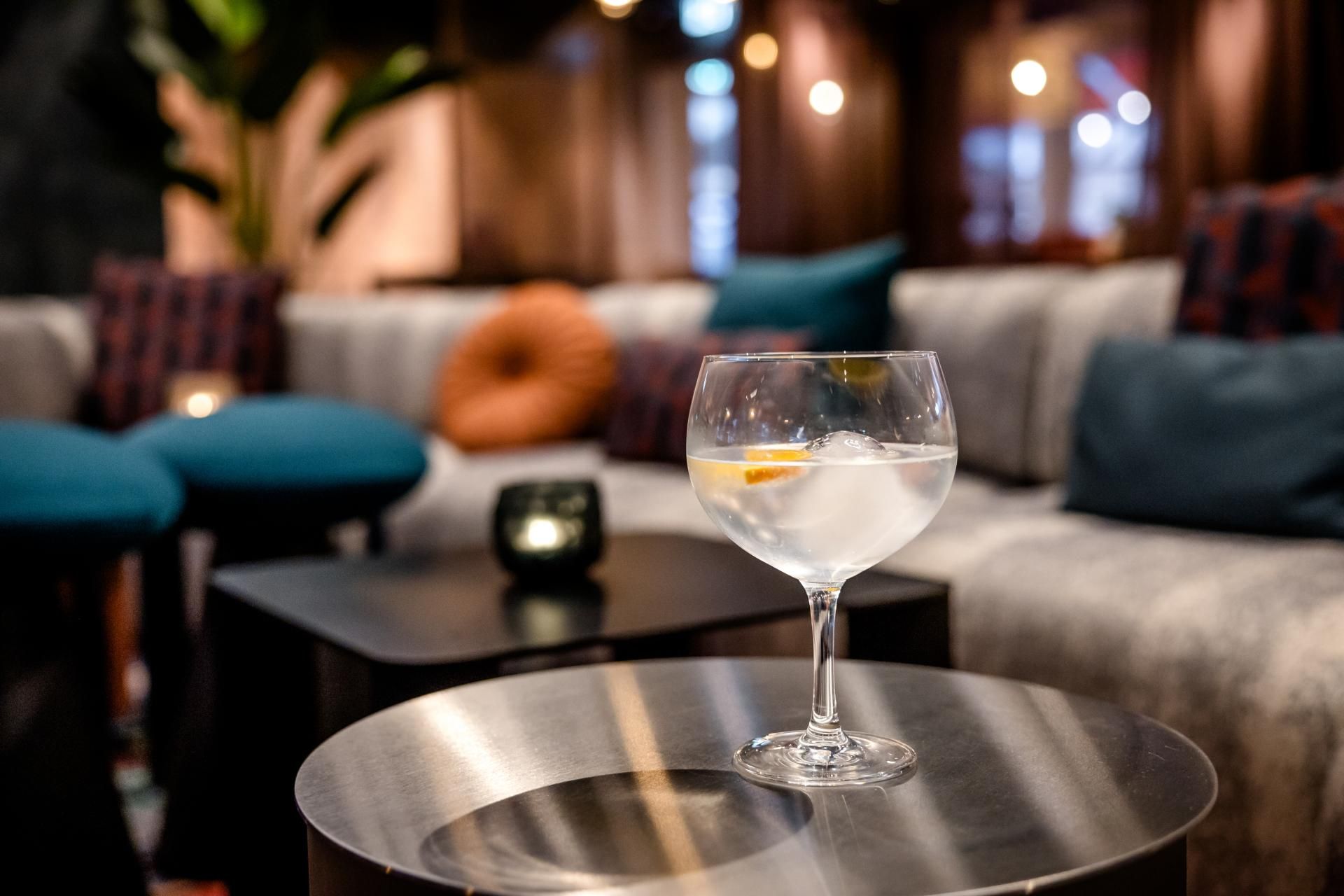 Ein elegantes Lounge-Setting mit gemütlichen Sofas und farbigen Kissen, im Vordergrund ein Cocktailglas mit einem Stück Zitrone auf einem runden Metalltisch, stimmungsvolle warmweiße Beleuchtung im Hintergrund.