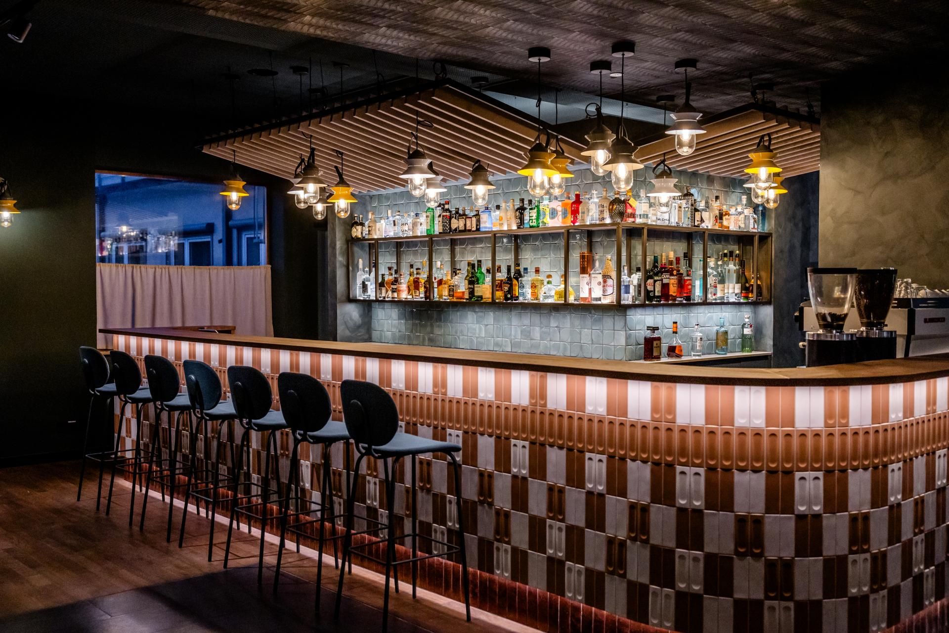 Moderne Hotelbar mit stilvoll beleuchtetem Tresen, schwarzen Barhockern und einer Auswahl an Spirituosen im Hintergrund, die eine elegante und einladende Atmosphäre für Abendunterhaltung bietet.