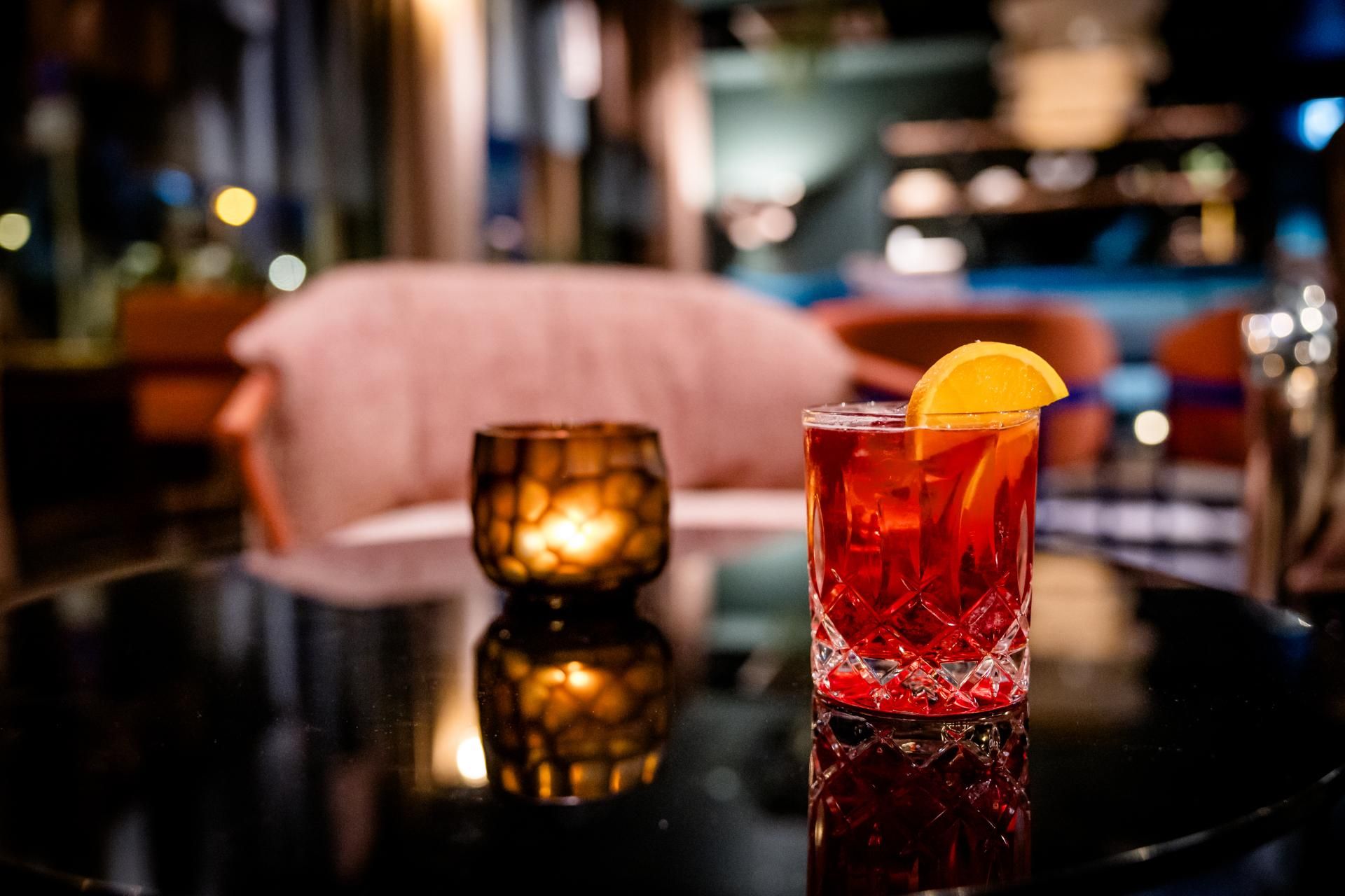 Eleganter Cocktail mit Orangenscheibe auf schwarzem, glänzendem Tisch neben einer atmosphärischen Kerze in gemütlicher Bar-Umgebung bei gedämpftem Licht.