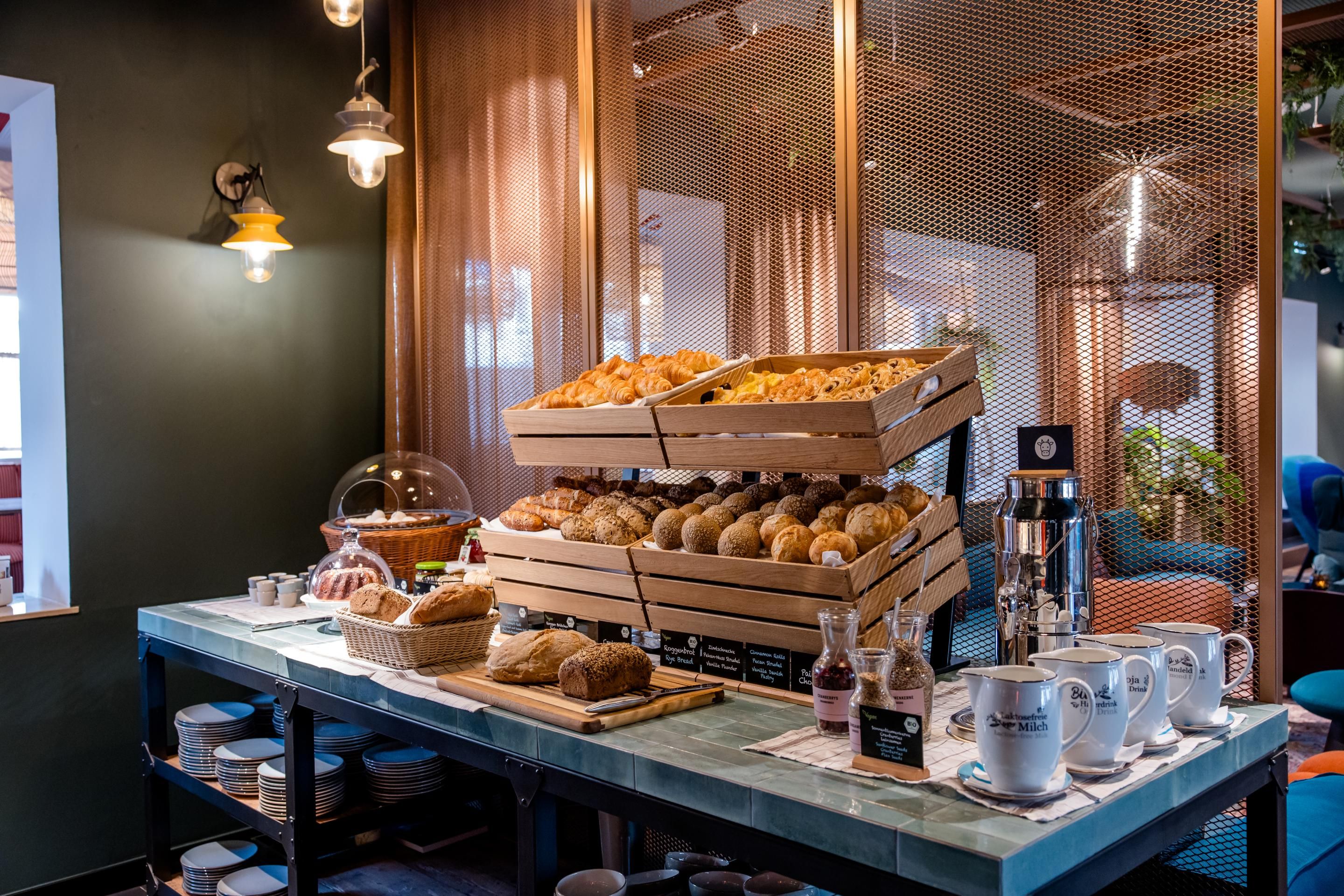 Frisch belegtes Frühstücksbuffet mit verschiedenen Brötchen, Brot, Croissants und Honig, liebevoll präsentiert in rustikalen Holzkisten, begleitet von Tassen und Kaffeekannen in einem gemütlichen, warm beleuchteten Ambiente.