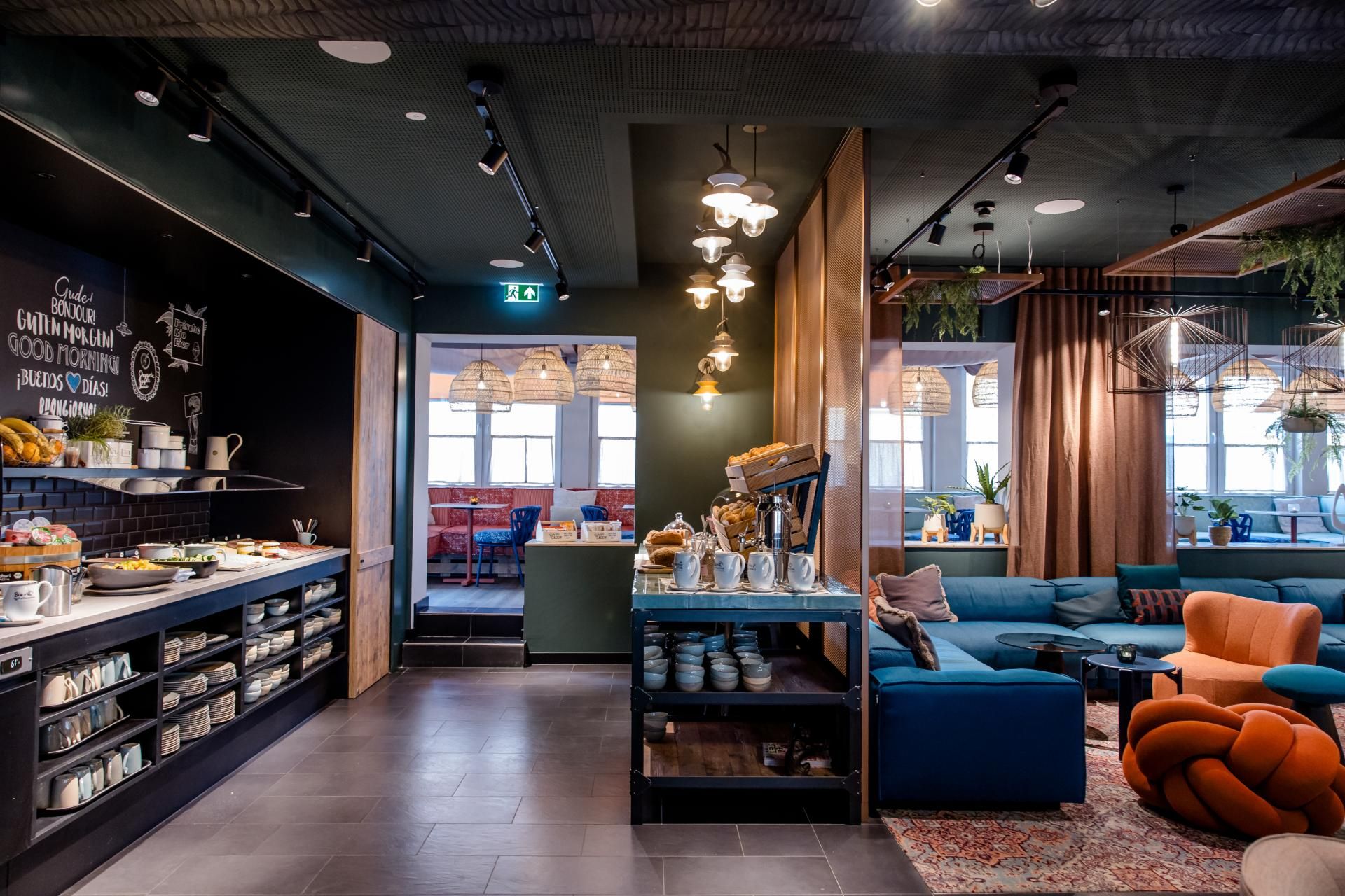 Modern eingerichteter Hotel-Loungebereich mit gemütlichen Sitzmöbeln, gedecktem Buffet und dekorativer Beleuchtung, der zum Entspannen und gemeinsamen Verweilen einlädt. Große Fenster lassen viel Tageslicht herein.