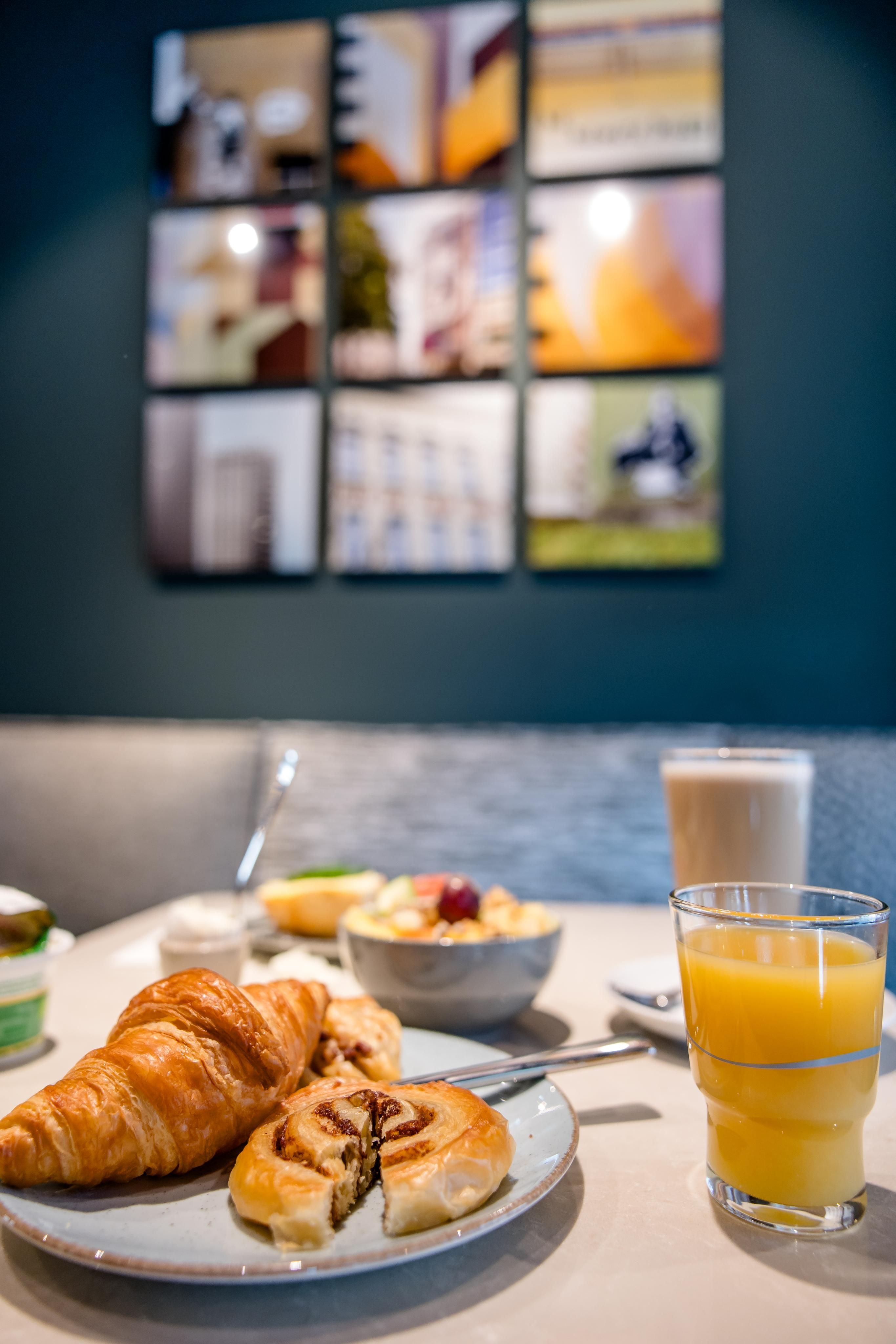Frühstückstisch mit Croissant, Schokoladenteilchen, Müsli, Glas Orangensaft und Latte Macchiato in entspannter Café-Atmosphäre.