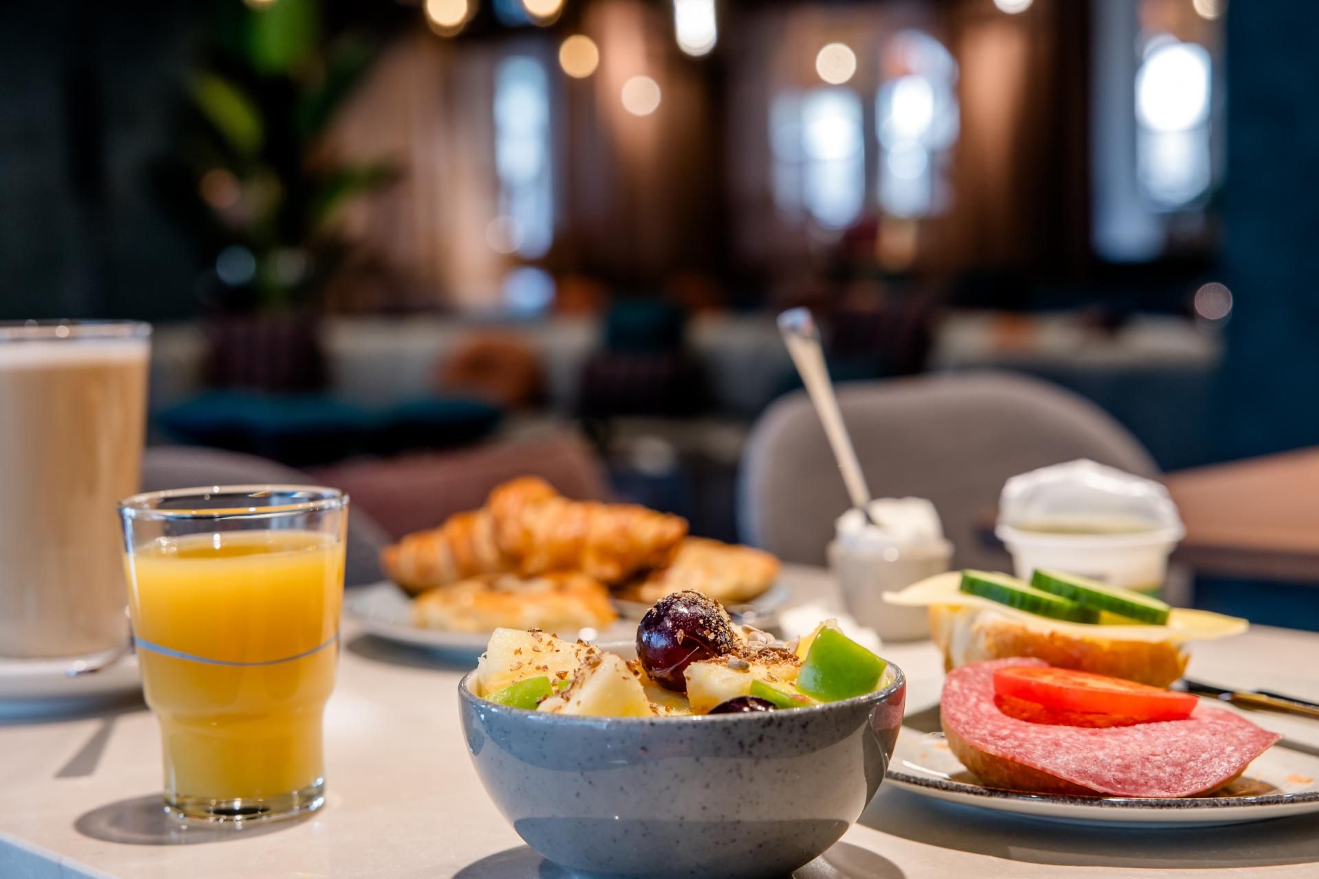 Frühstückstisch mit frischem Orangensaft, Müsli, Croissants und belegten Broten in gemütlicher Café-Atmosphäre.